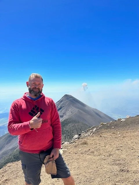 A man with a beard and red hoodie giving a thumbs-up on a mountain trail with a volcanic mountain and smoke in the background under a clear blue sky.