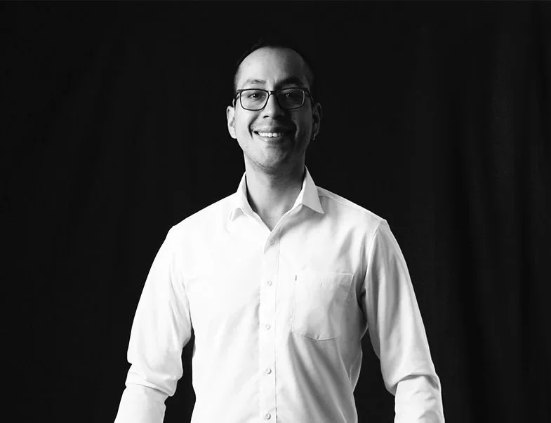 Hombre con gafas y camisa blanca sonriendo, fondo oscuro.