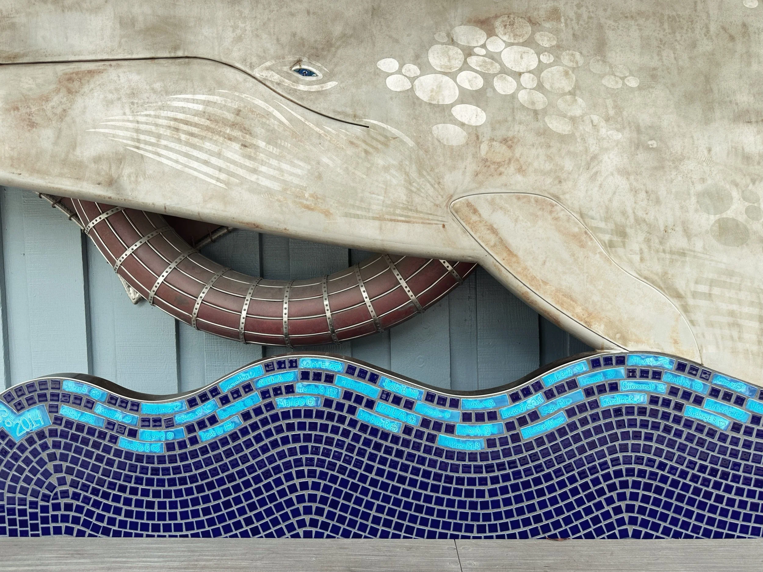 A close-up of a colorful mosaic with names written in blue tiles at the bottom, and a large white whale sculpture with a water spout on a textured wall in the background.