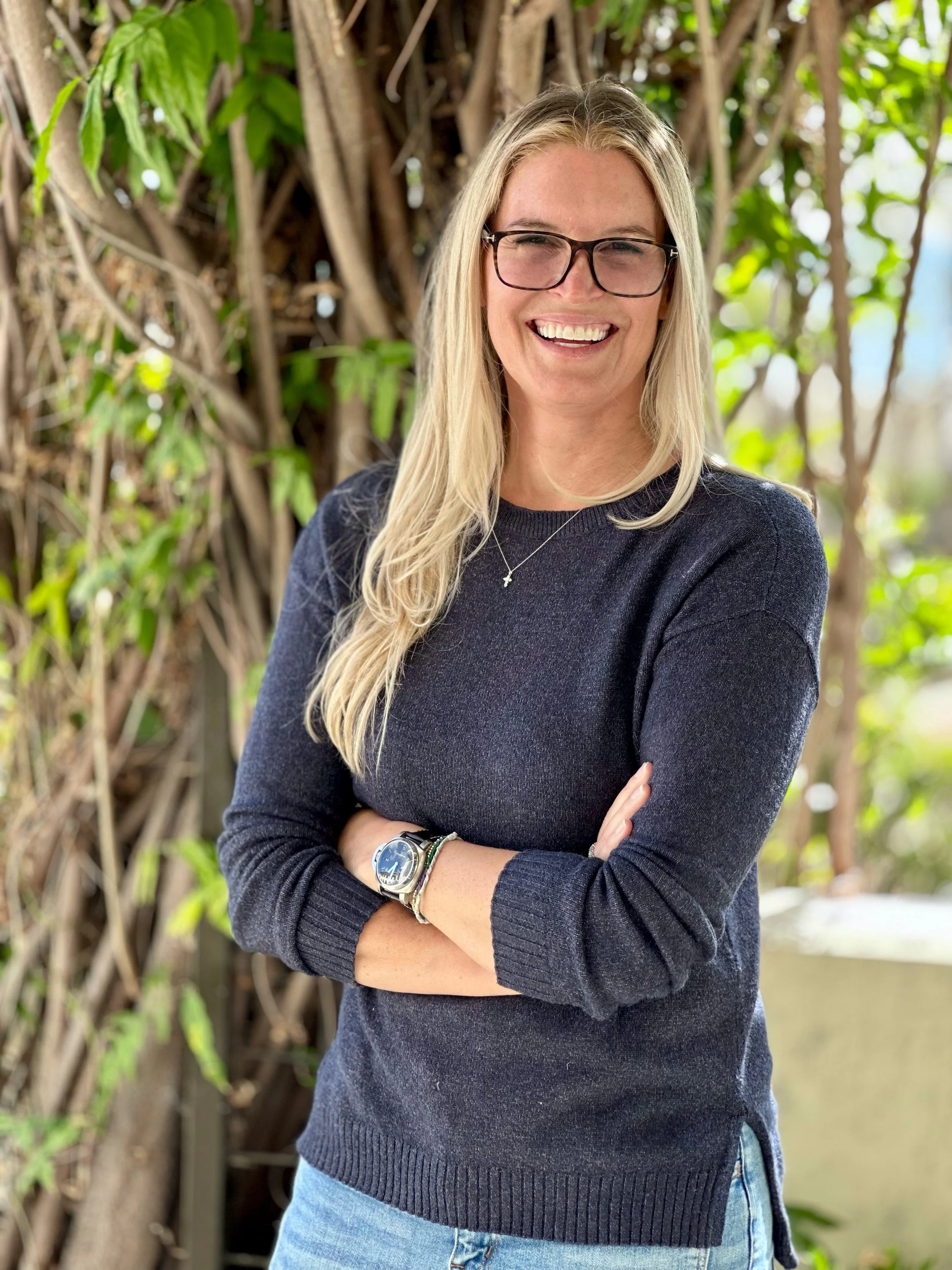 A woman with long blonde hair, glasses, and a big smile, standing outdoors with crossed arms, wearing a dark sweater and jeans, with greenery and tree branches in the background.