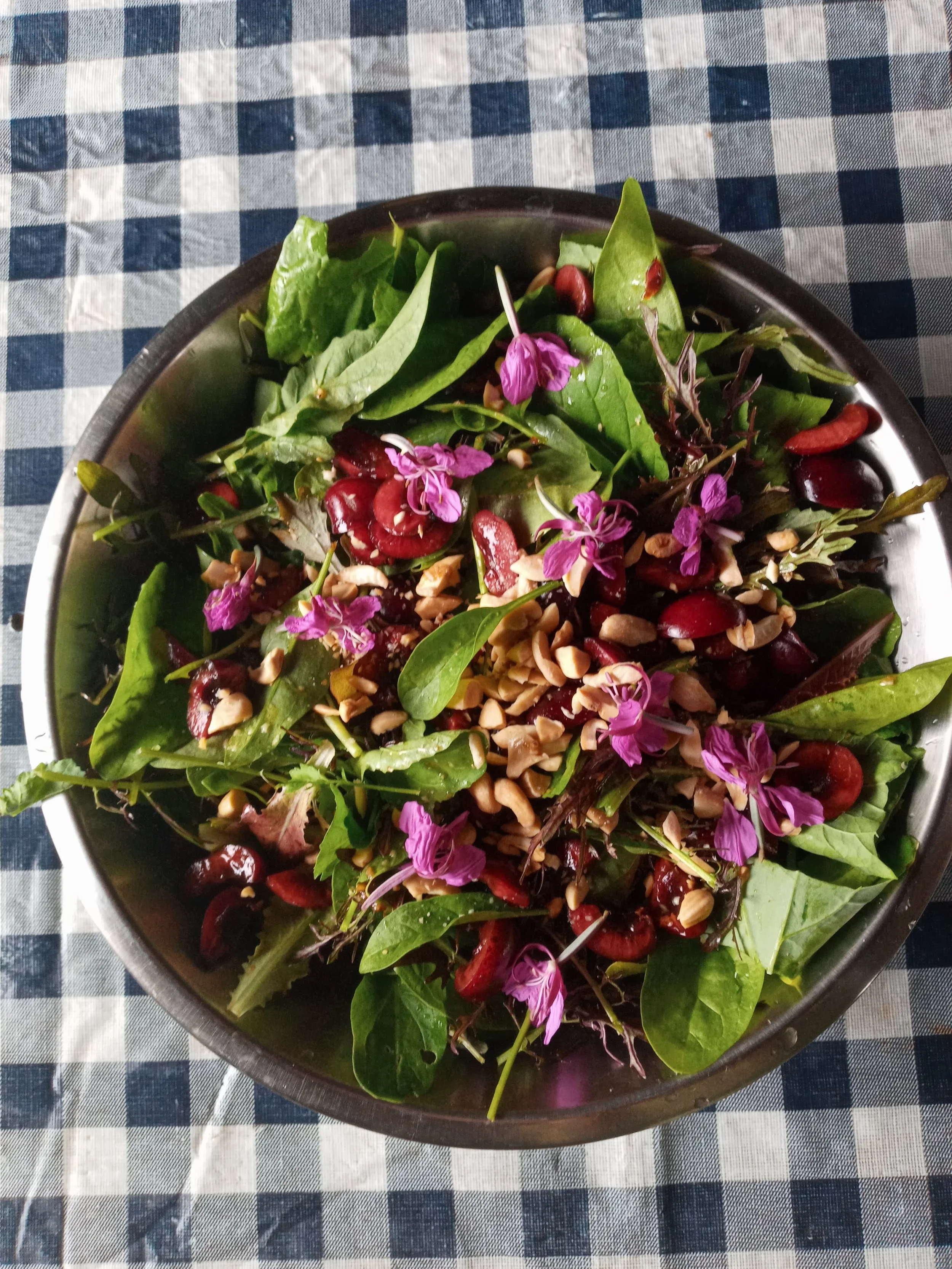 A bowl of mixed salad with greens, cherry tomatoes, chopped nuts, and pink edible flowers on a checkered tablecloth.