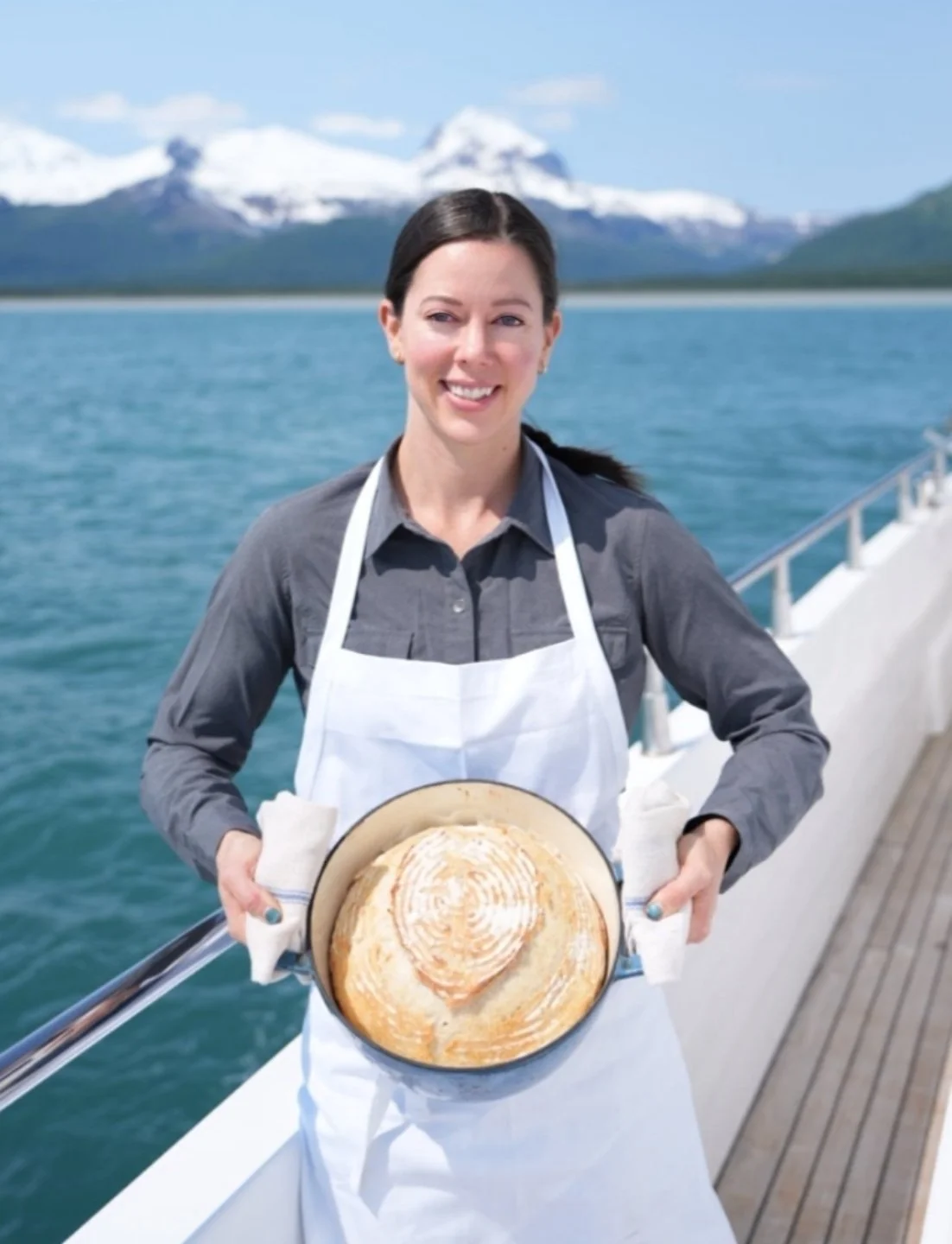 Woman on a boat holding a freshly baked sourdough bread in a pan with a scenic mountain and water background.