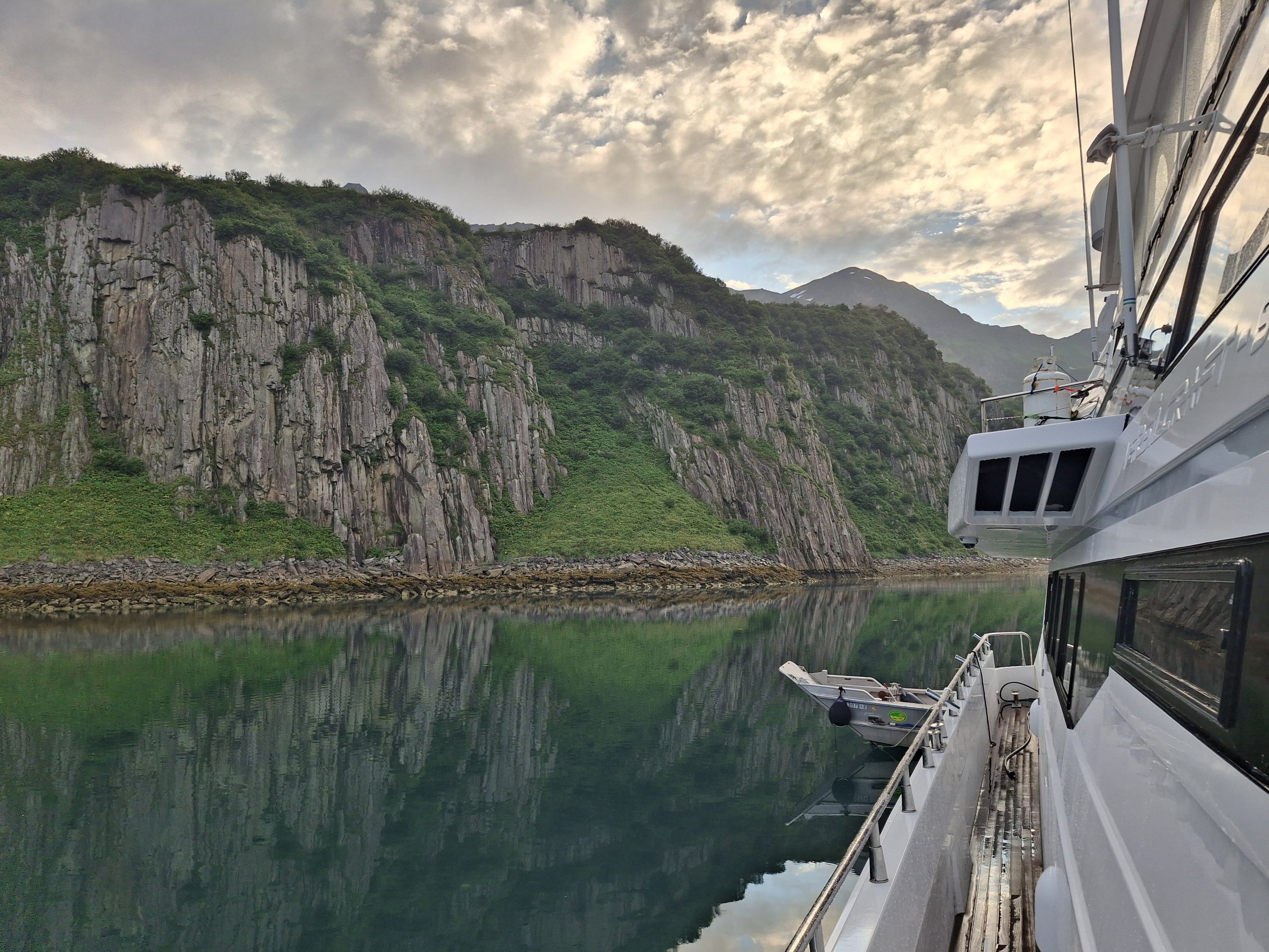 A large white motor yacht docked in a calm, green water body with rocky, green mountains and a cloudy sky in the background.