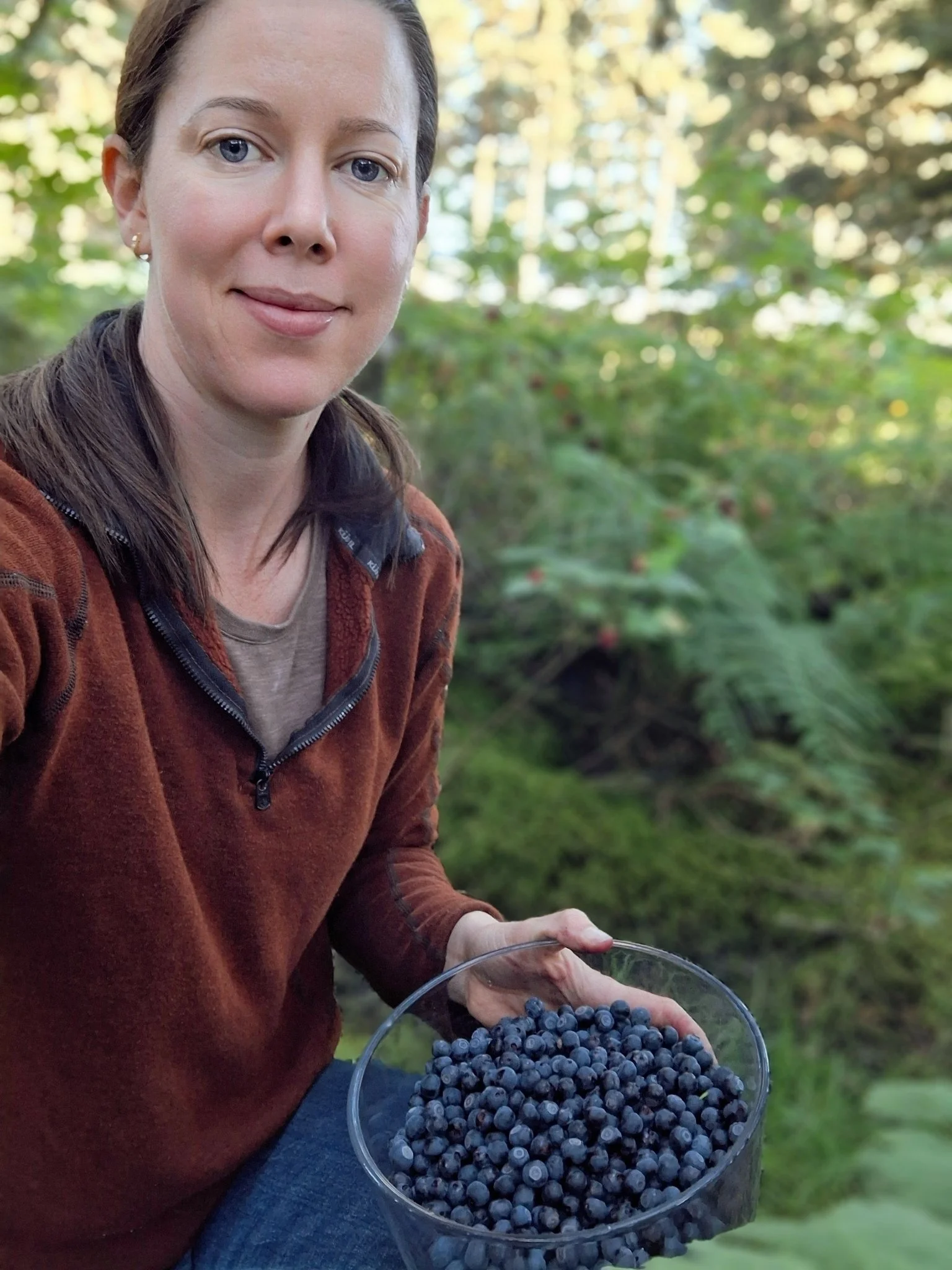 A woman with blue eyes and long brown hair, wearing a reddish-brown fleece jacket and sitting outdoors, is holding a glass bowl filled with fresh blueberries.