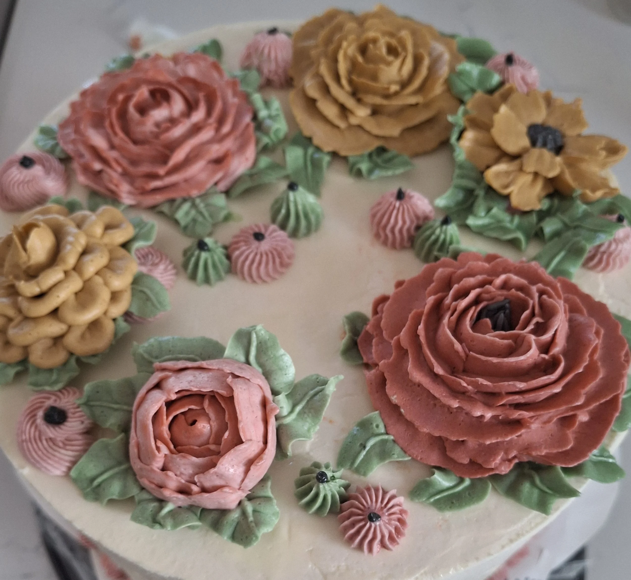 Decorative cake with pink, yellow, and green floral icing, featuring roses and leaves, topped with small black dots.