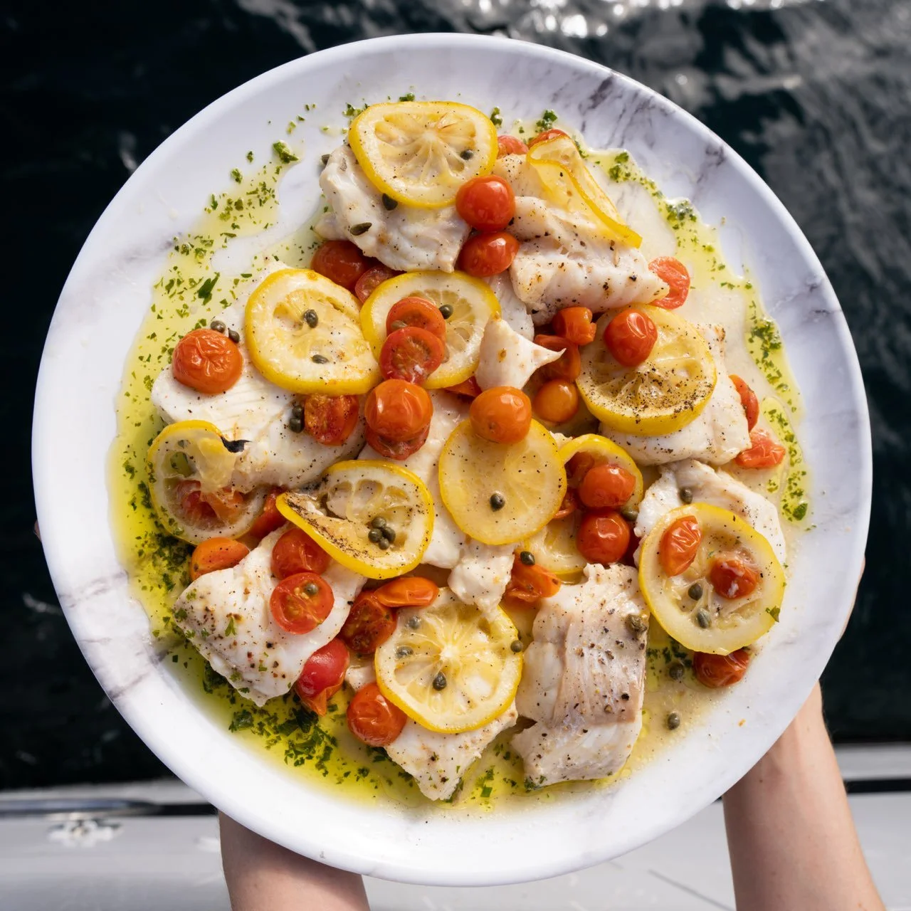 Fresh white fish fillets topped with lemon slices, cherry tomatoes, and capers, drizzled with olive oil and sprinkled with herbs.