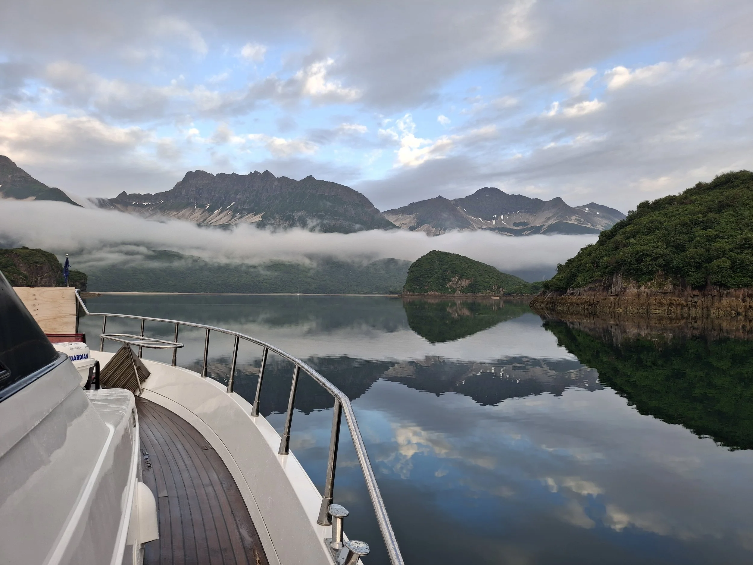 View from a boat showing calm water, green hills, and mountains in the background with partly cloudy sky and some fog or low clouds over the mountains.
