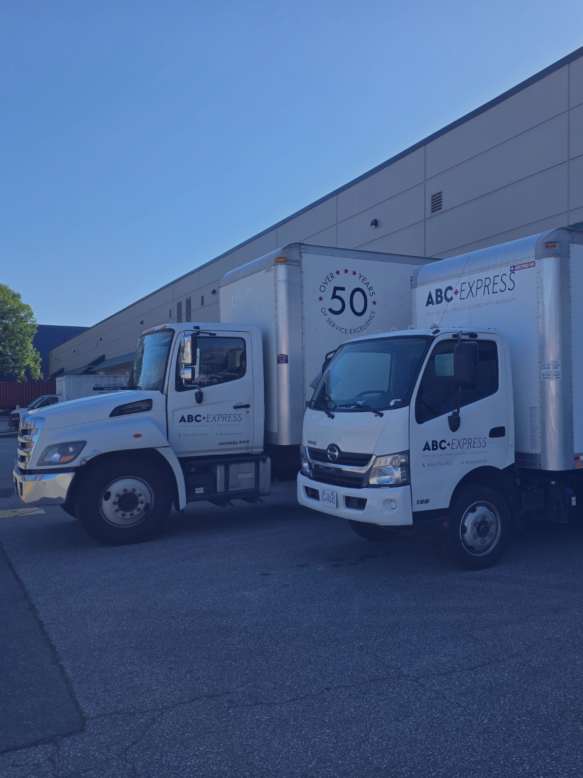 Two ABC Express delivery trucks parked side by side in front of a large beige industrial building under a clear blue sky.
