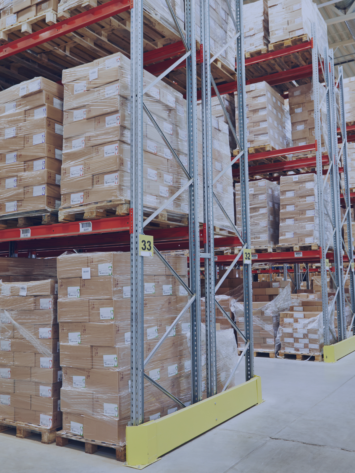 Warehouse aisle with shelves stocked with boxed wooden planks wrapped in plastic on pallets, labeled with barcodes and numbers.