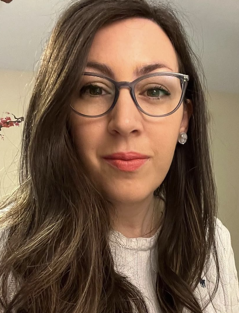 A woman with long brown hair, wearing glasses, pearl earrings, and a light-colored sweater.