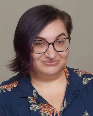 A woman with short dark hair and glasses, wearing a blue floral shirt, looking at the camera with a slight smile.