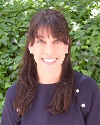 A woman with long brown hair and bangs smiling outdoors with green foliage in the background.