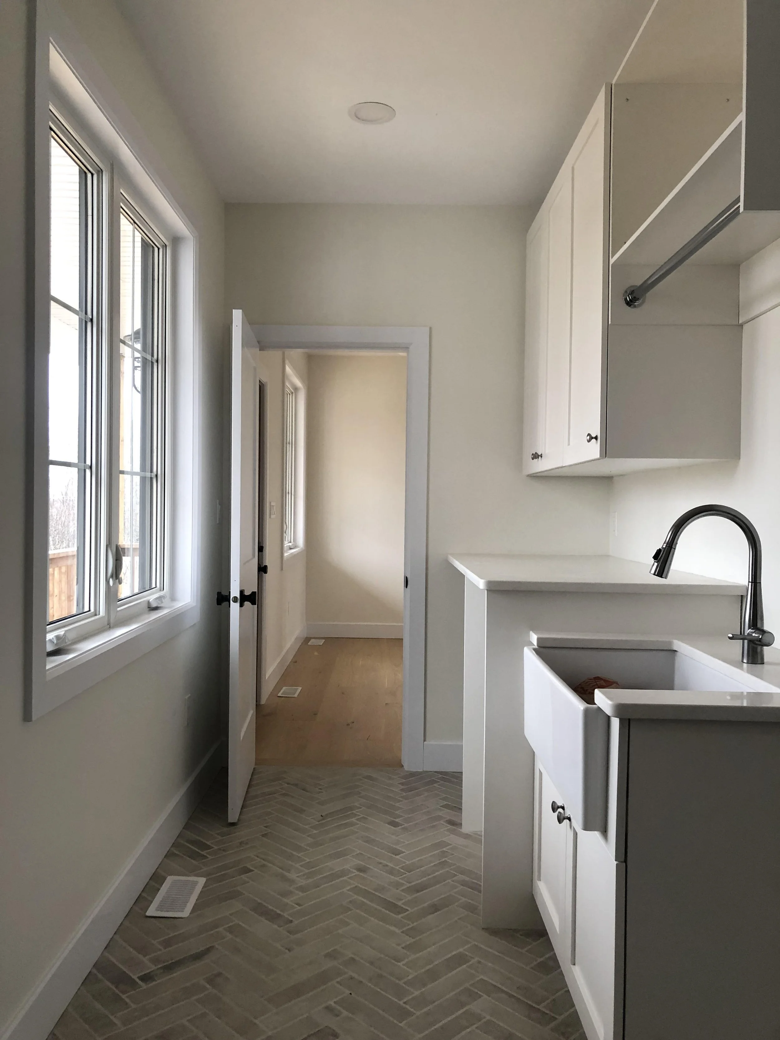 Interior of a kitchen with large window, white cabinets, a sink with a black faucet, and a door leading outside, with beige tile flooring.