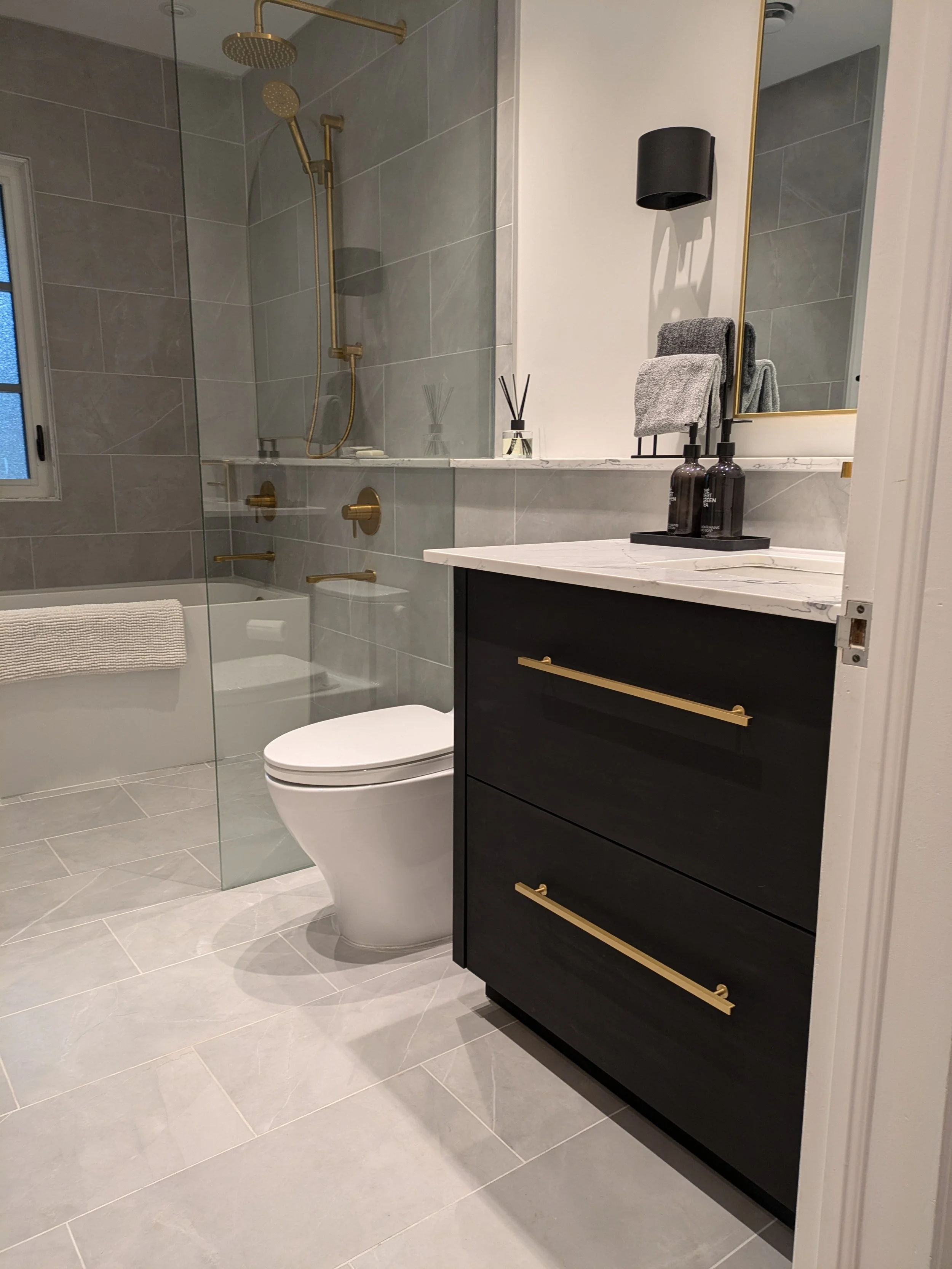 Modern bathroom with a walk-in shower featuring gold fixtures, a bathroom vanity with a white marble countertop, black cabinetry with gold handles, and a large mirror. Towels and bottles are on the vanity, and a window with frosted glass is in the shower area.