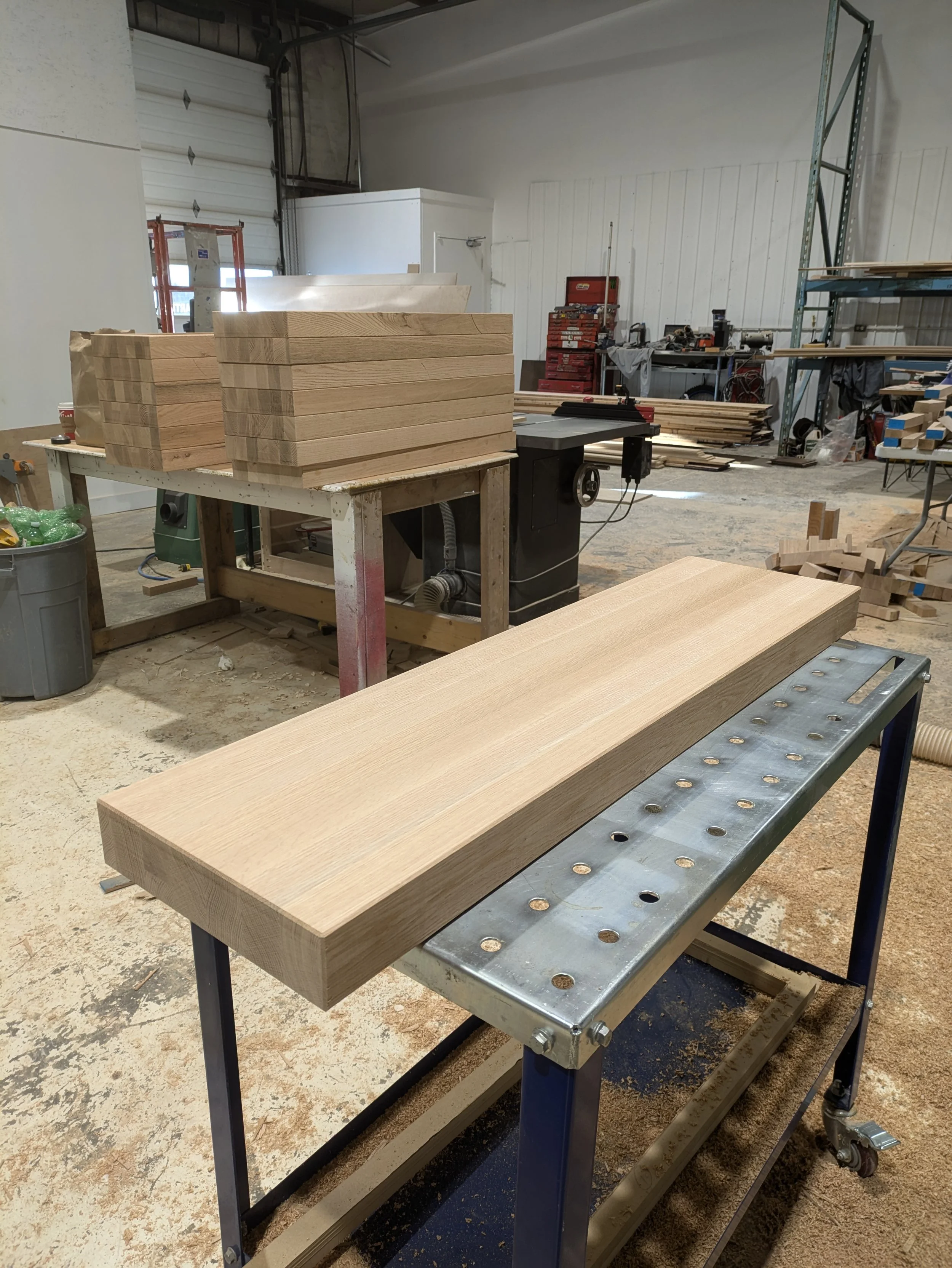 A woodworking workshop with a workbench holding several stacks of cut wooden planks, a table saw, and various tools and materials scattered in the background.
