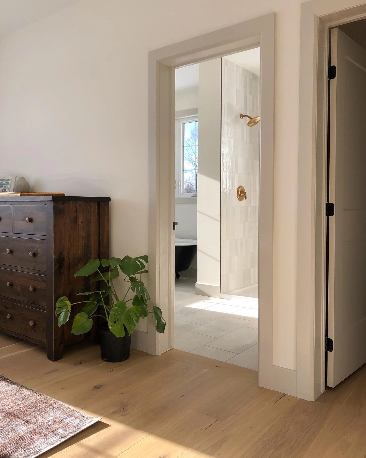 View of a bathroom with a glass shower door, shower fixtures, and window seen through an open doorway from a room with light-colored walls, wooden floor, a dark wood dresser, and a potted plant. Custom trim and doors. New construction.