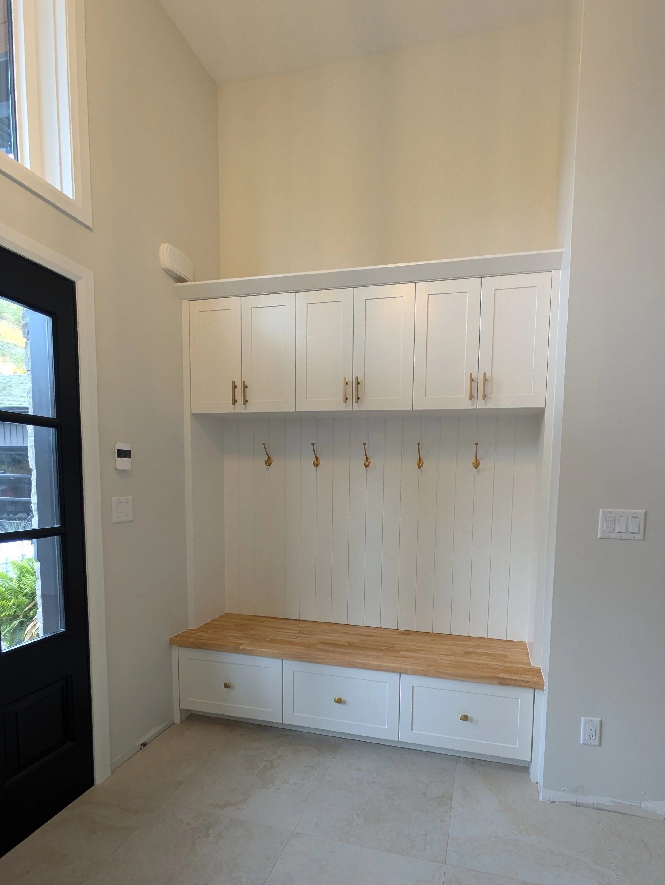 Interior view of a modern hallway with white built-in cabinets, a wooden bench, and hooks for hanging items, adjacent to a black-framed glass door with natural light coming in.