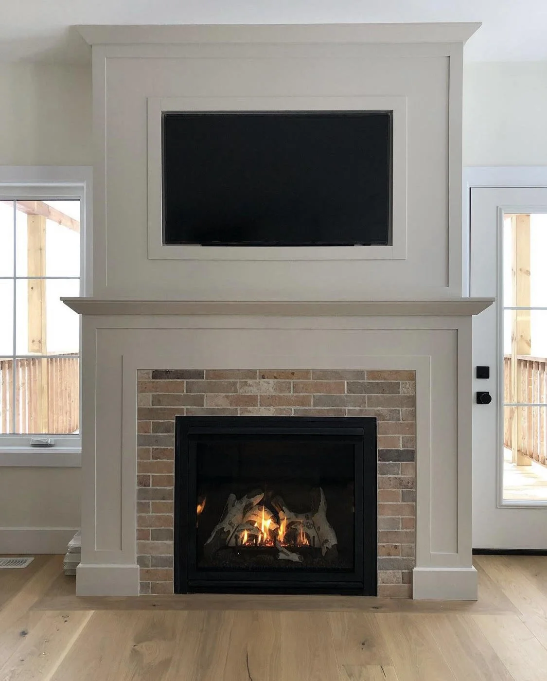 Living room fireplace with brick surround, mantel, and a mounted flat-screen TV above. Windows on both sides, showing an outdoor balcony area.
