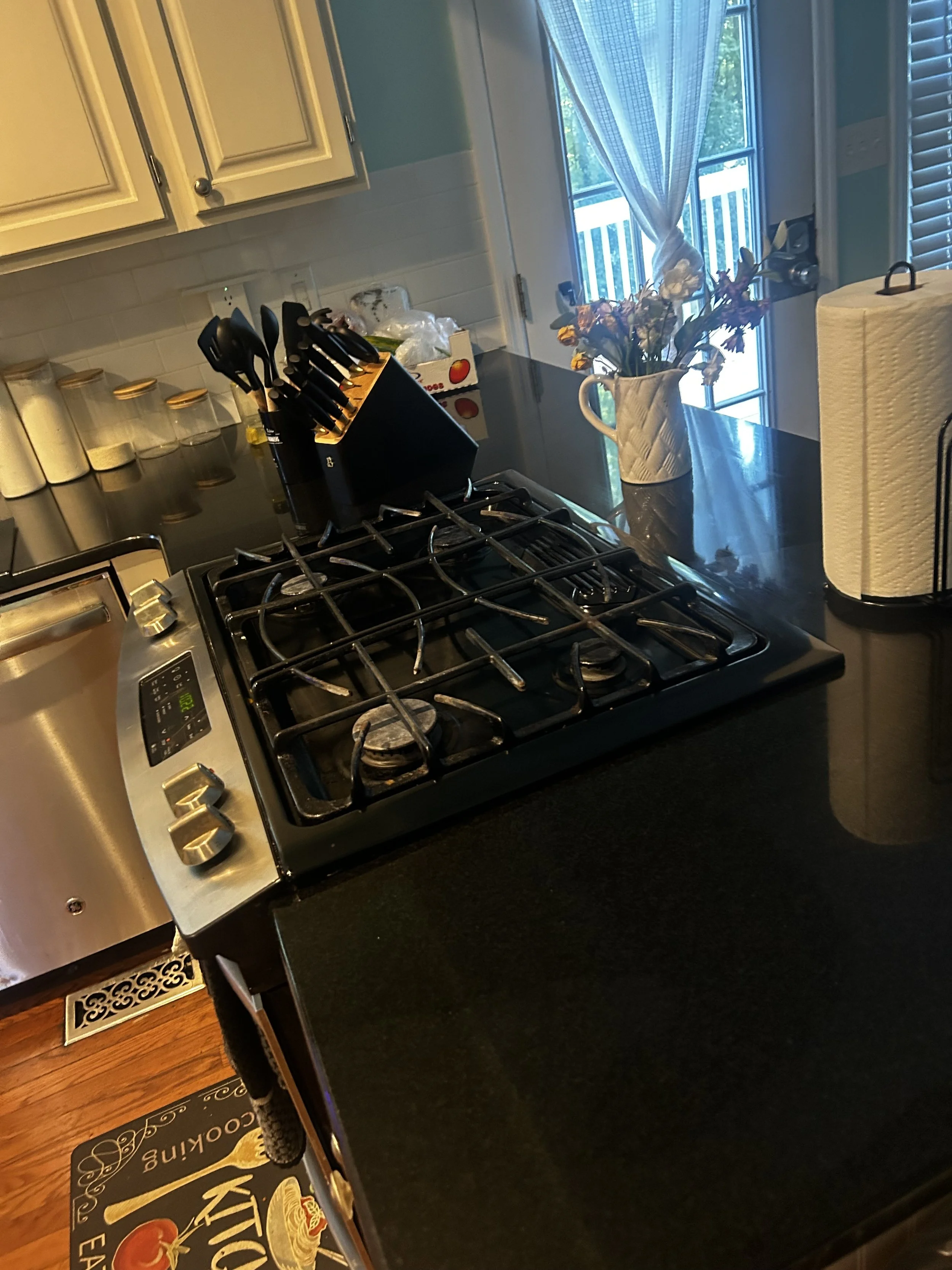 Kitchen countertop with a stovetop, a knife block, a paper towel roll, a decorative flower vase, and a window with curtains.