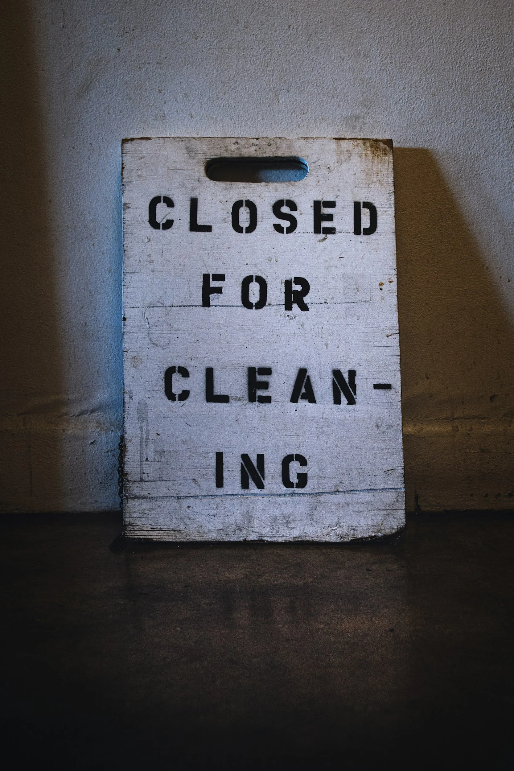A weathered wooden sign with black stencil text reading 'CLOSED FOR CLEANING' is leaning against a wall. The sign has a handle cutout at the top and appears worn with some scratches and chipping paint.