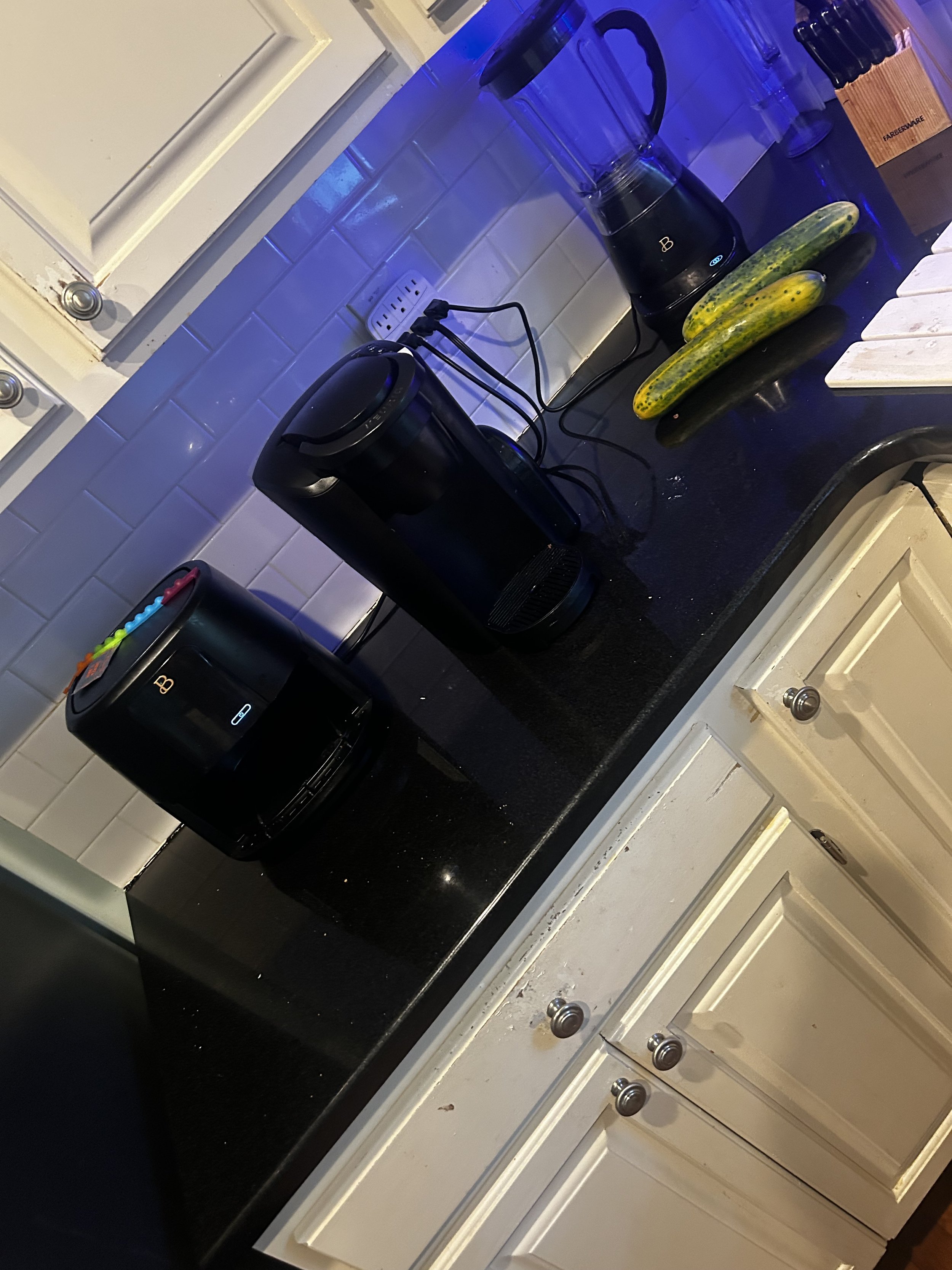 Kitchen countertop with a black coffee maker, a coffee grinder, a cucumber, a yellow cucumber, some clear glasses, a box of beer bottles, and a white chopping board. The kitchen has white cabinets and a white tile backsplash.