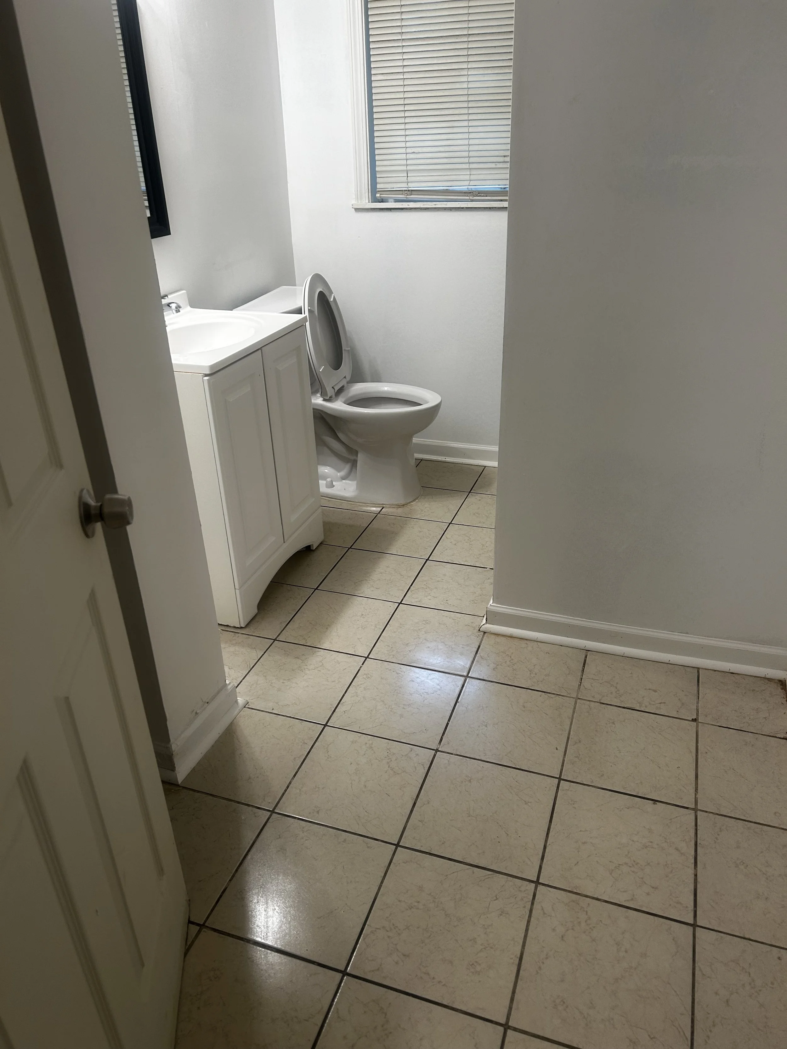 A small bathroom with beige tiled floor, a white cabinet with a sink, a toilet with the lid open, and a window with closed blinds.