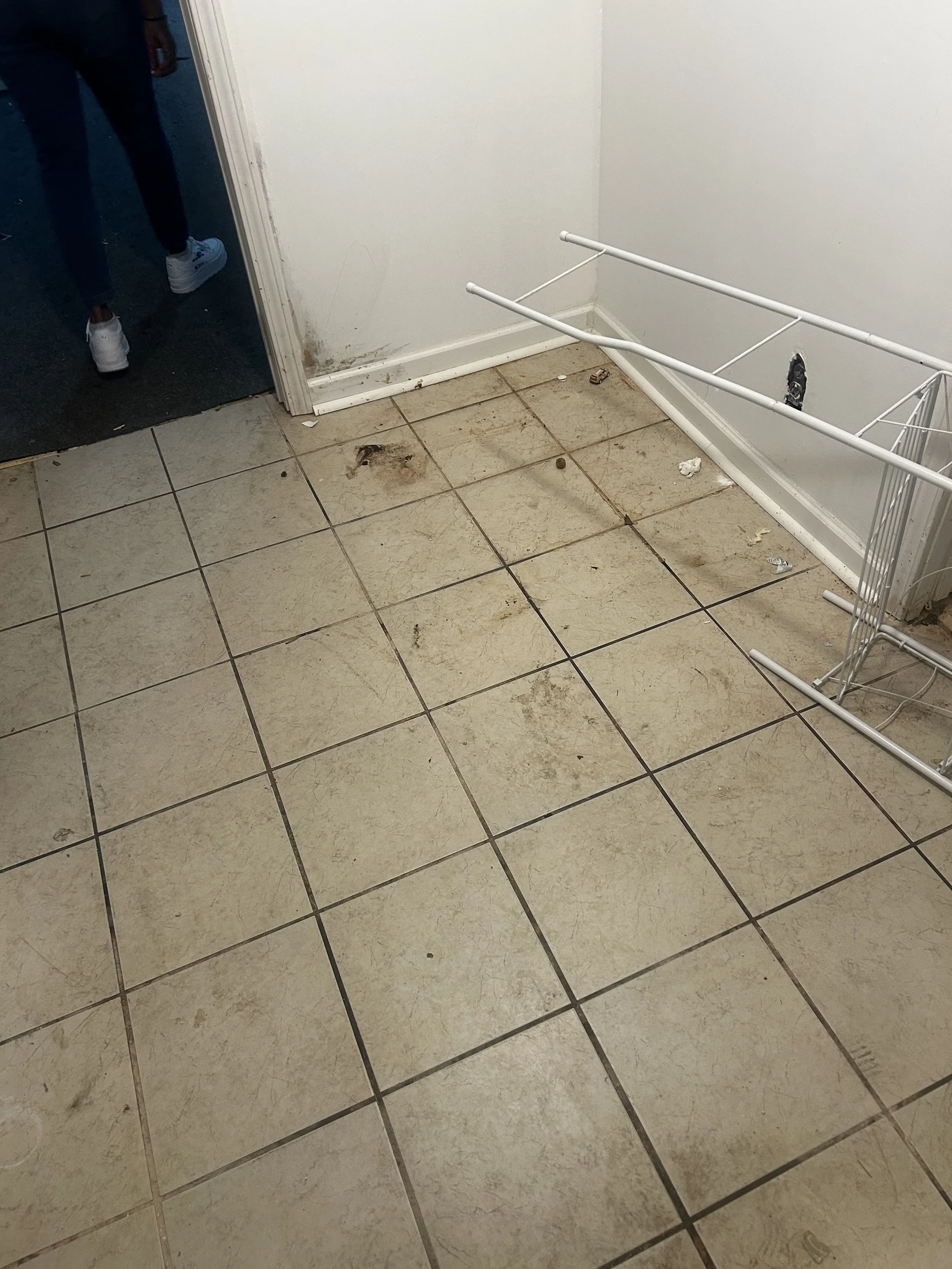 Dirty and stained tile floor with scattered trash, an empty white metal shelf, and a wall with an outlet and some damage.