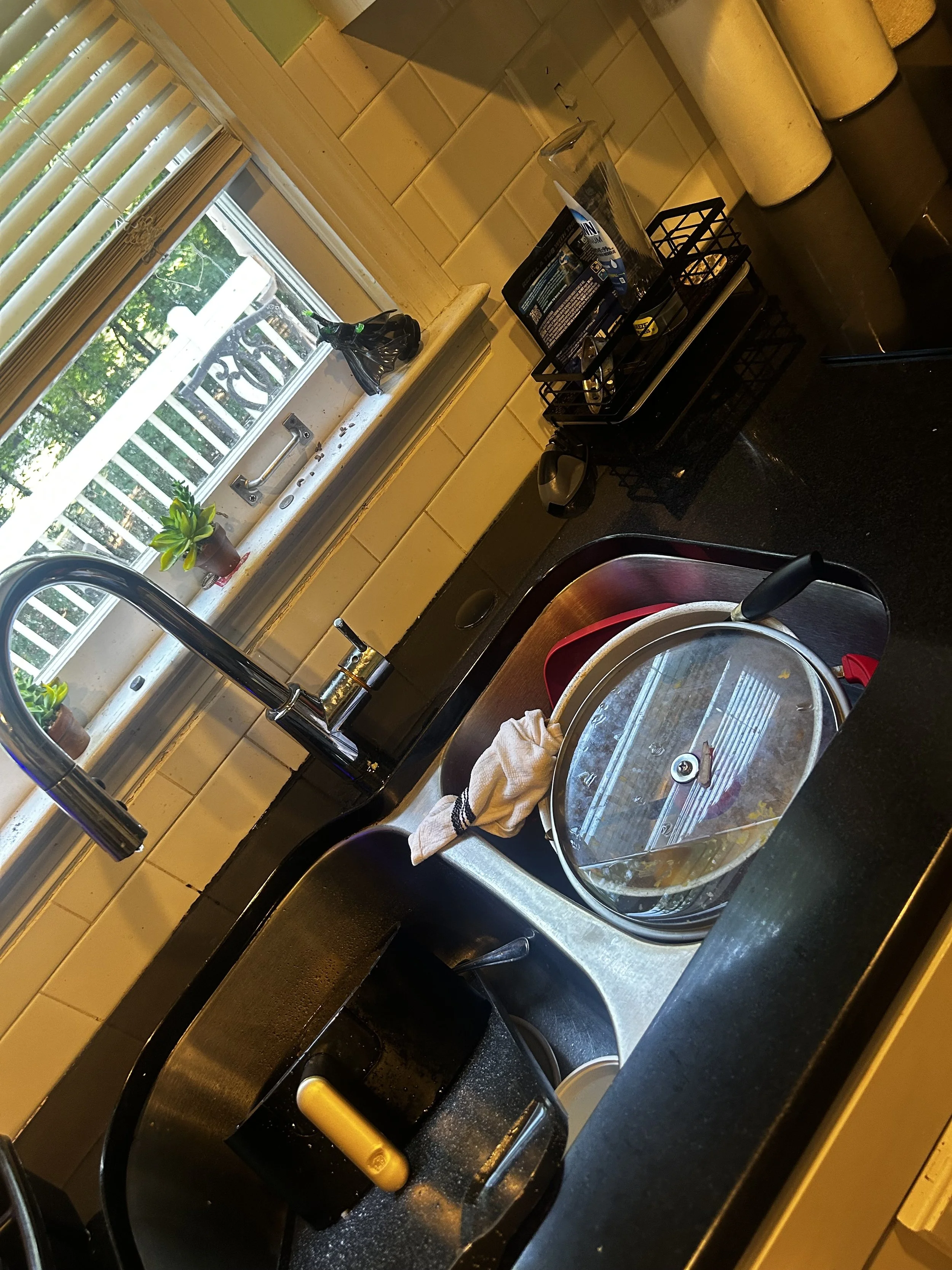 A kitchen countertop with a sink, a squeegee, a towel, a dish drying rack with utensils and snacks, a plastic cup, and a window with blinds and a small potted plant.