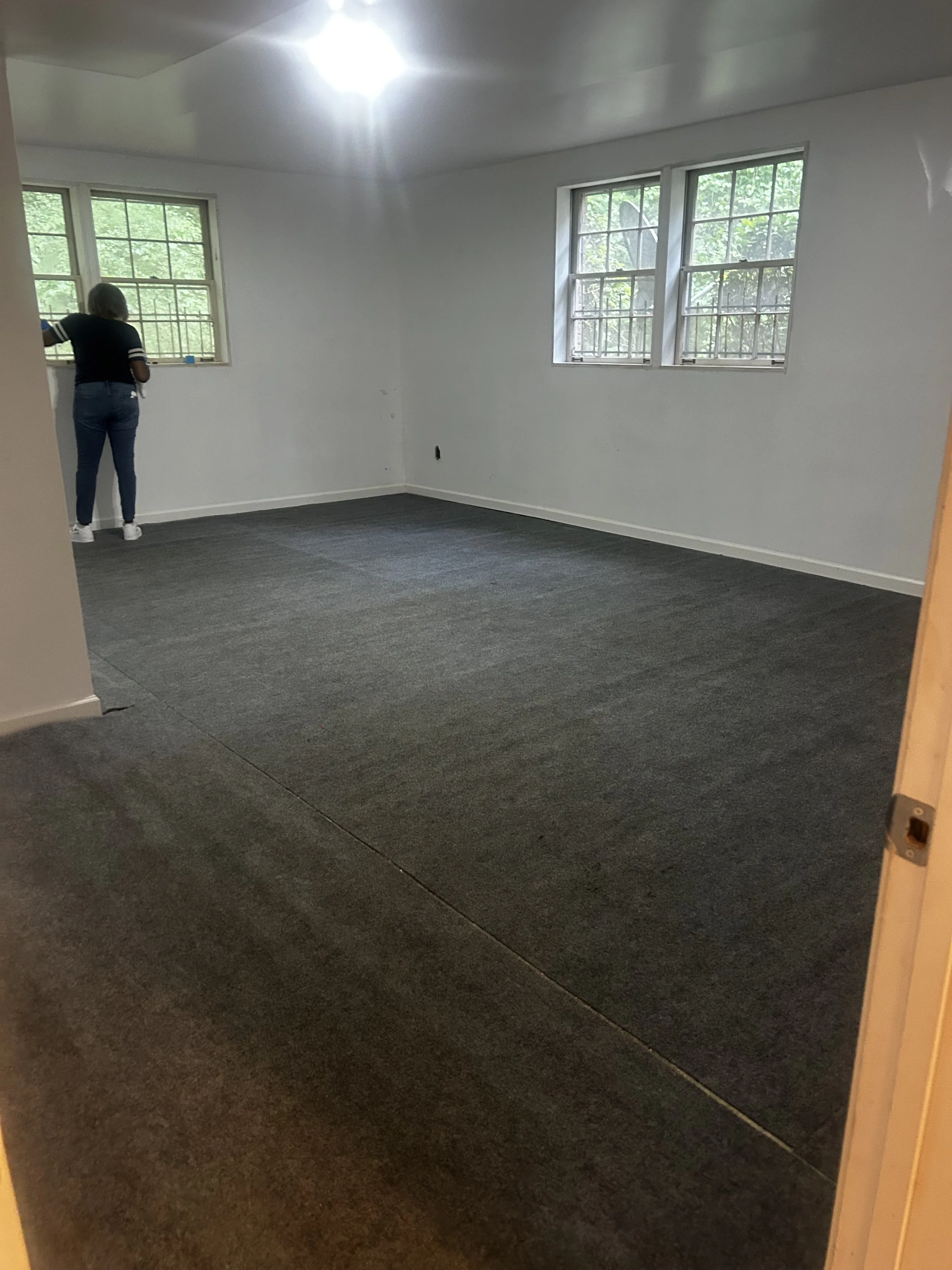 An empty room with white walls, three windows, a person with gray hair wearing a black shirt and blue jeans looking out the window, and a gray carpeted floor.