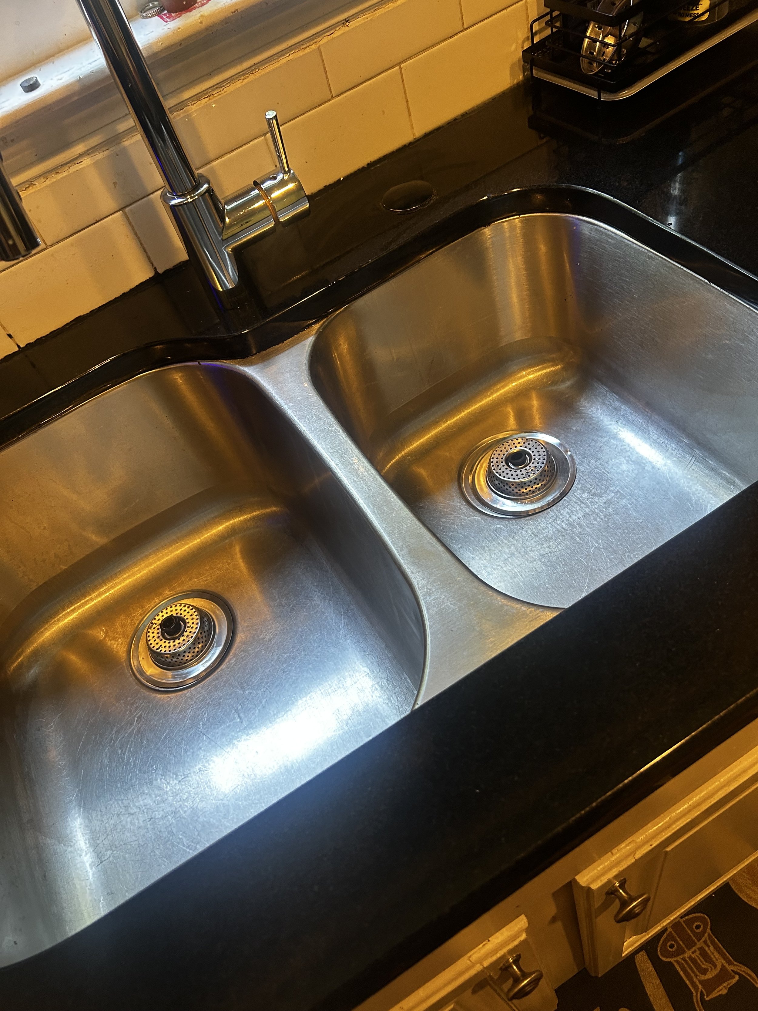 A stainless steel double kitchen sink located under a window, with a modern chrome faucet, black countertop, a black dish drying rack in the corner, and a tiled backsplash.
