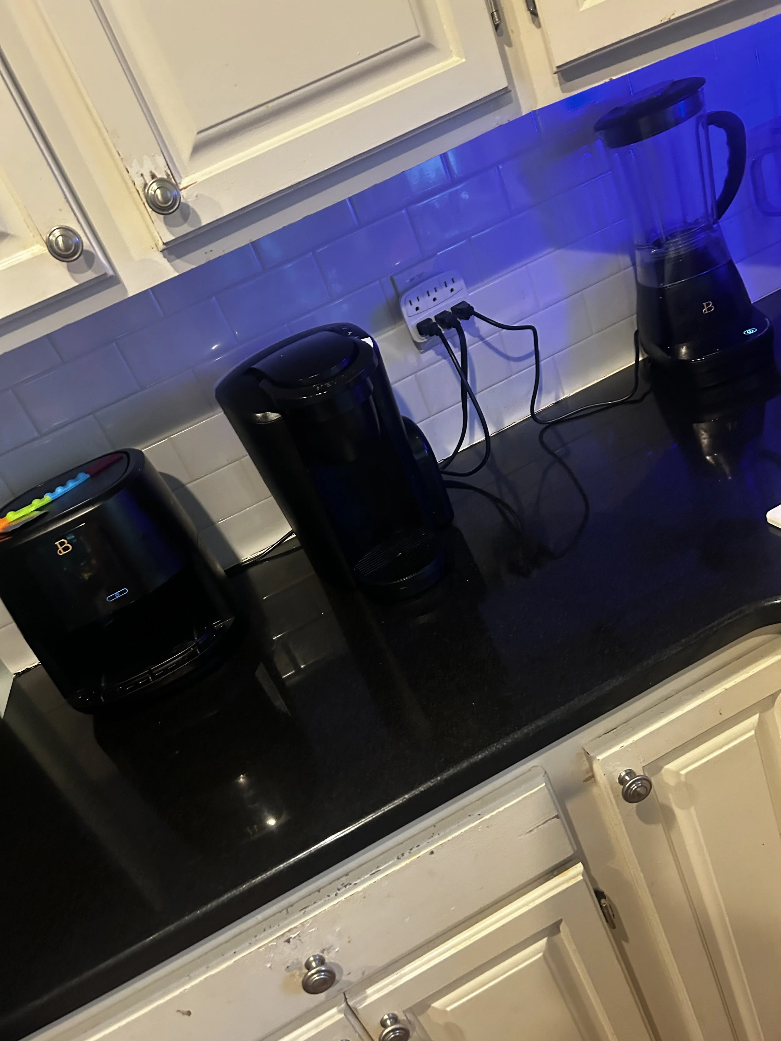 Kitchen countertop with black coffee makers plugged into an outlet and a glass blender with a blue light on the right side.