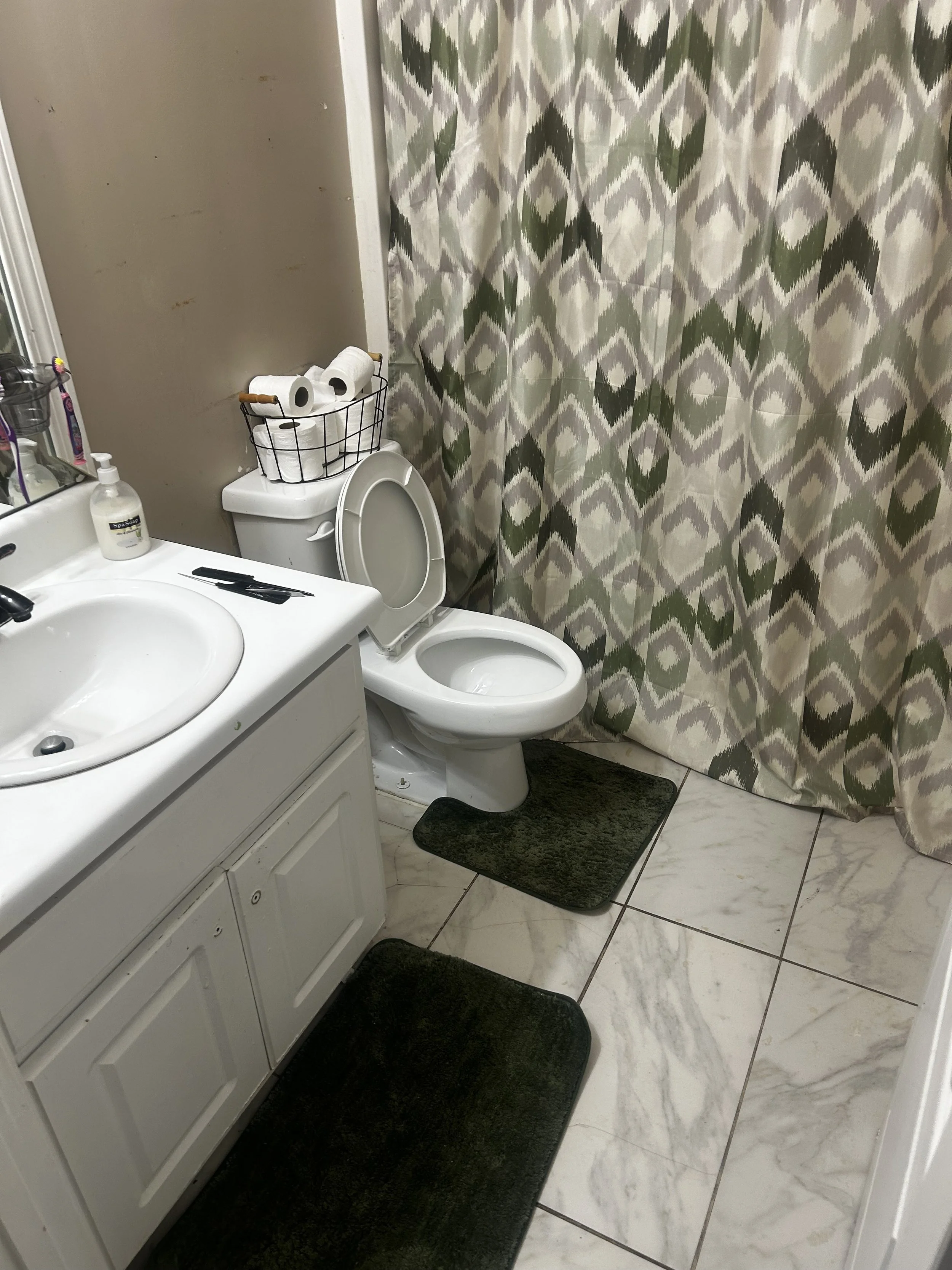 A bathroom with a sink, a cabinet below, a mirror, and a toilet with a wire basket on top holding toilet paper rolls. There are three dark green rugs on a white marble floor. A patterned shower curtain with green, beige, and gray designs covers the b