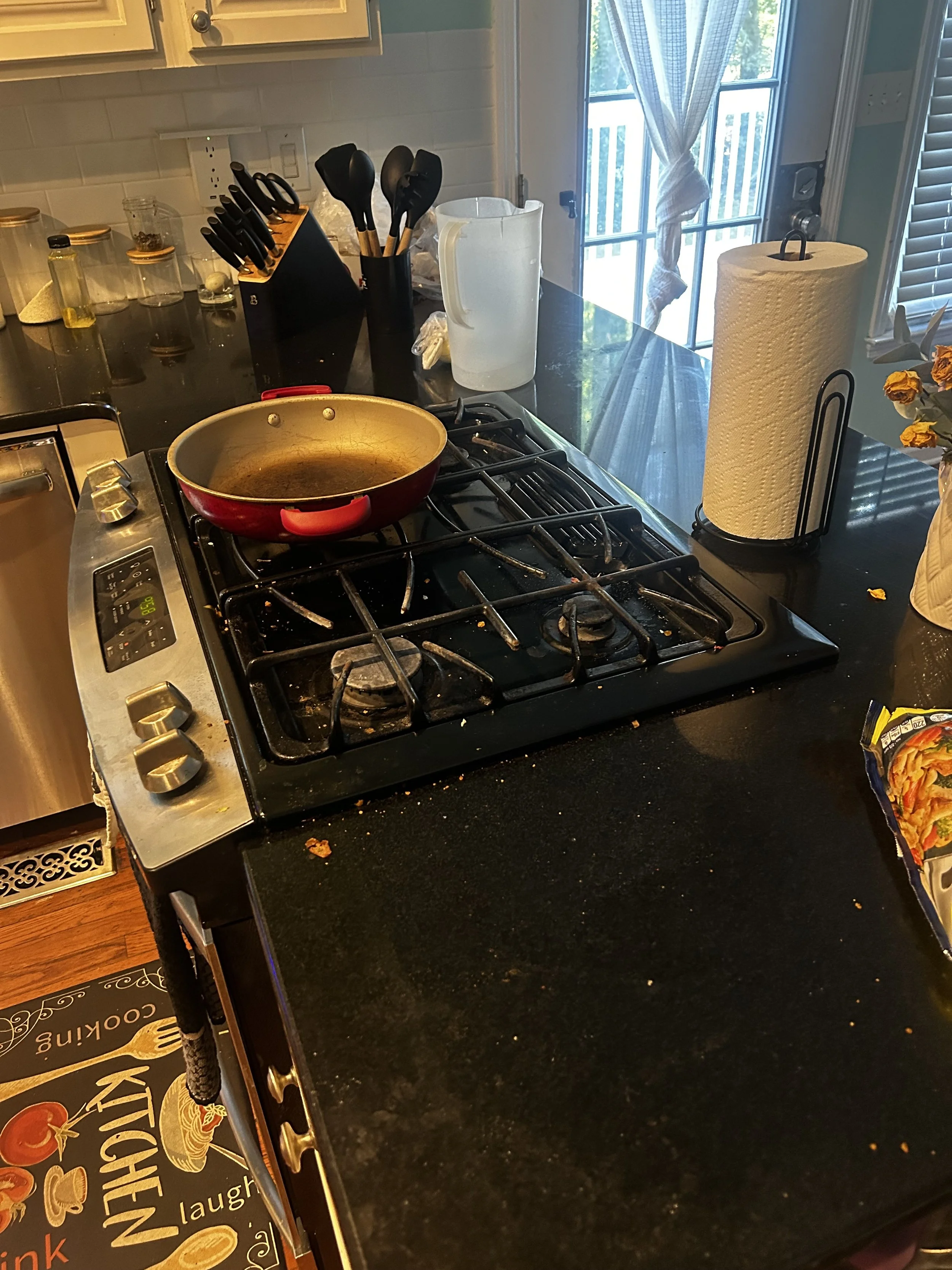 Kitchen counter with a red frying pan on a stove, kitchen utensils in a black holder, a roll of paper towels, and a water pitcher near a sliding door with a view outside.