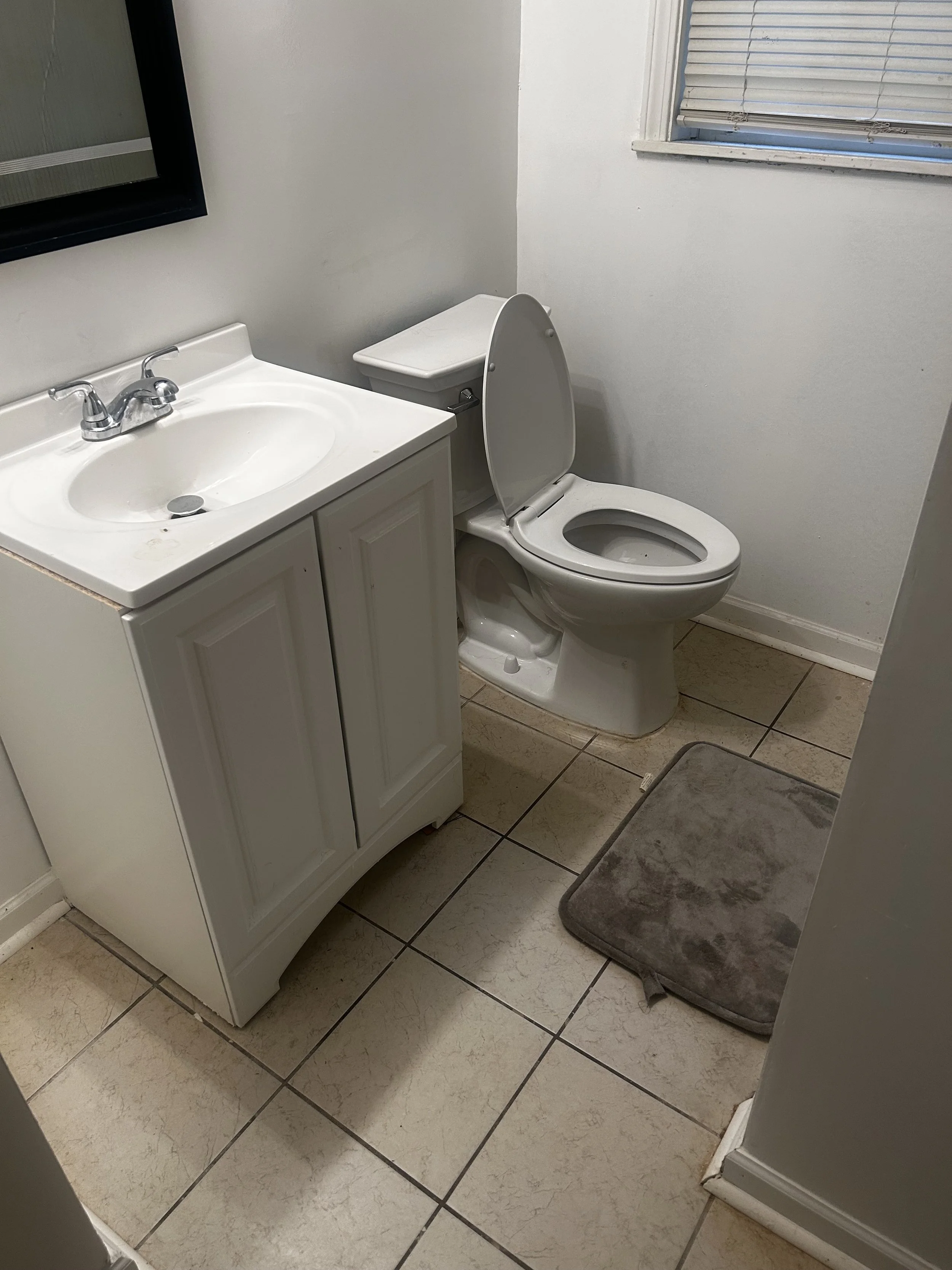 A small bathroom with a white vanity, sink, a mirror, a toilet with a lid, and a window with closed blinds. The floor is tiled and there is a gray rug in front of the toilet.