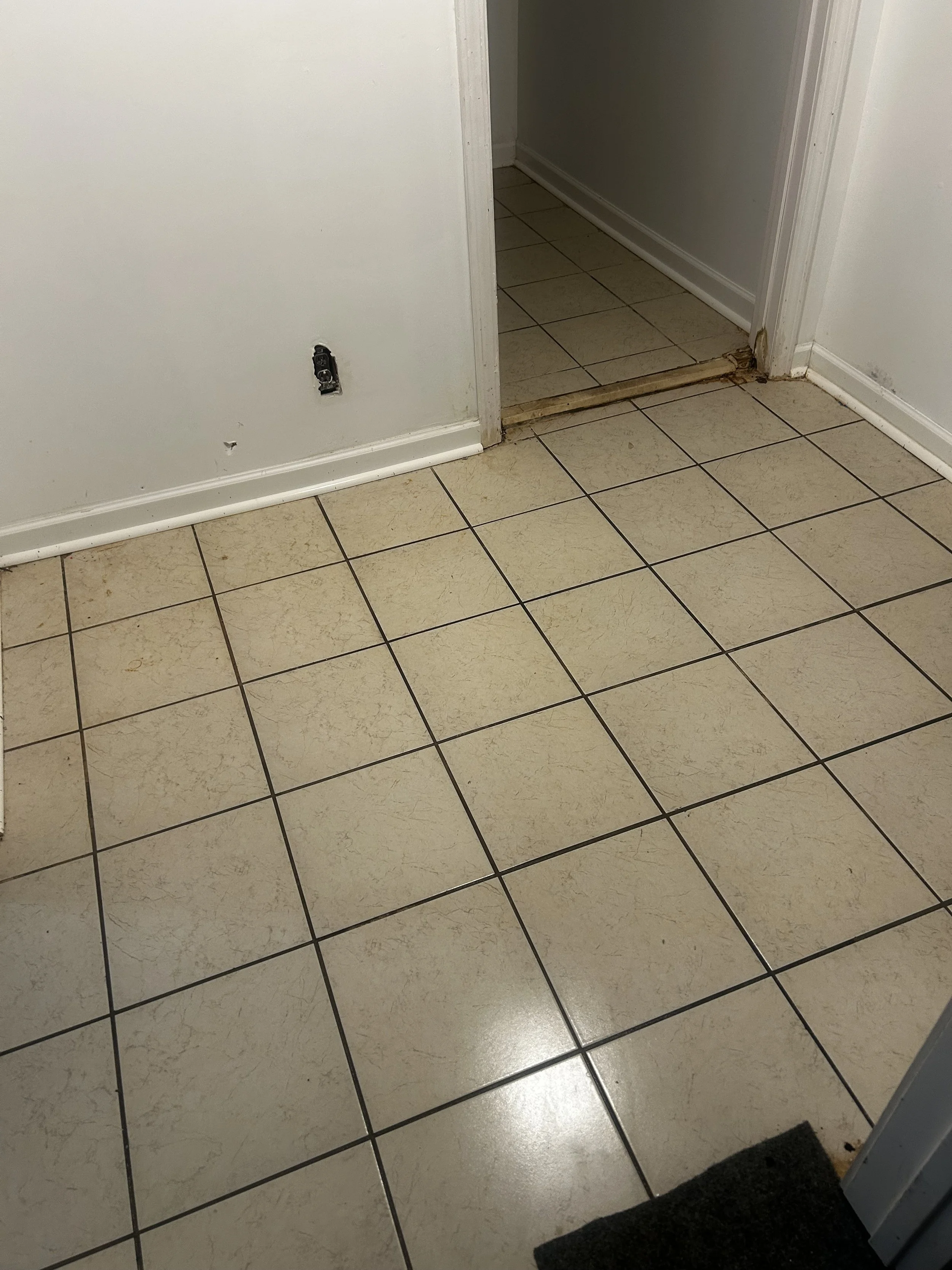 The image shows a section of an interior space with beige tiled flooring. An opening between two rooms reveals a portion of the adjacent room with similar tile flooring. The wall at the center has a loose electrical outlet cover, and there is visible