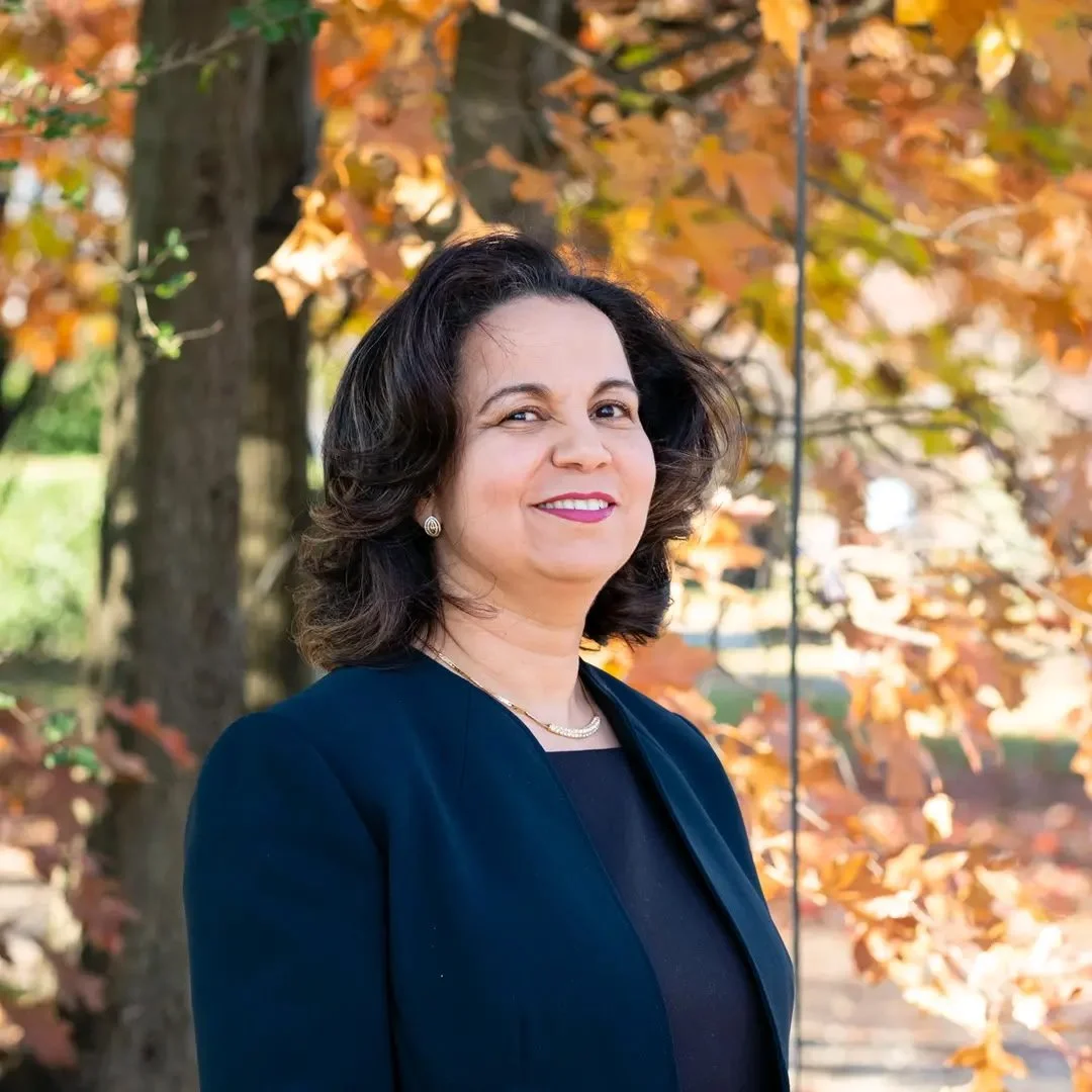 A woman with dark brown hair, wearing a navy blazer and a black top, standing outdoors in front of a tree with autumn-colored leaves, smiling at the camera.