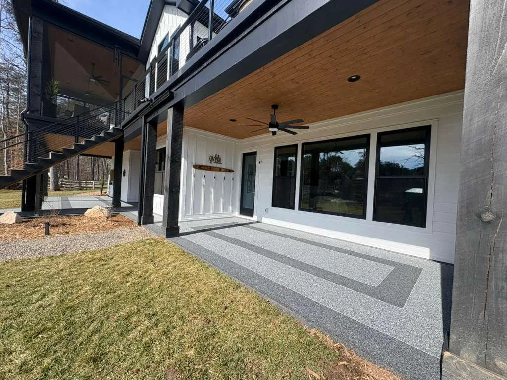 Back porch of a modern house with large windows, a ceiling fan, and a gray and white patterned concrete floor. There is a black staircase leading to the upper deck and some landscaping with rocks and mulch.