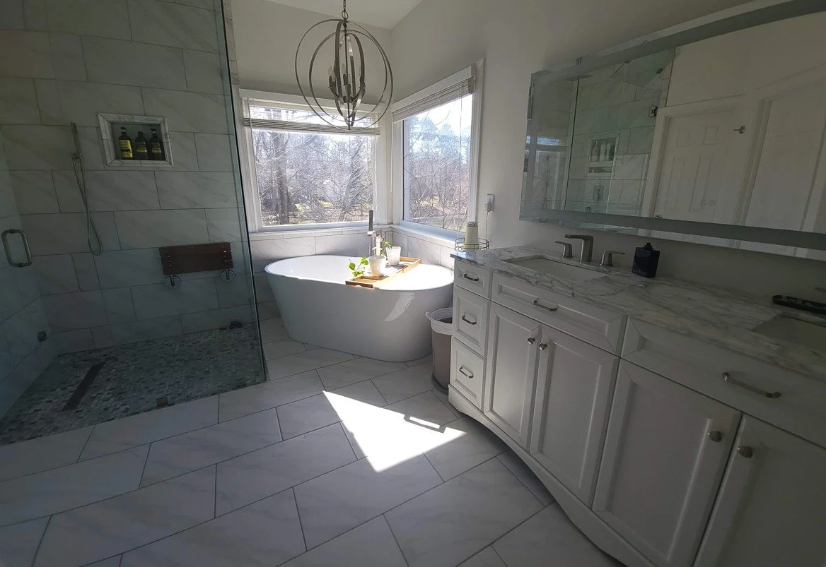 Bright bathroom with a large window, a white bathtub, a vanity with marble countertop, and a glass-enclosed shower.
