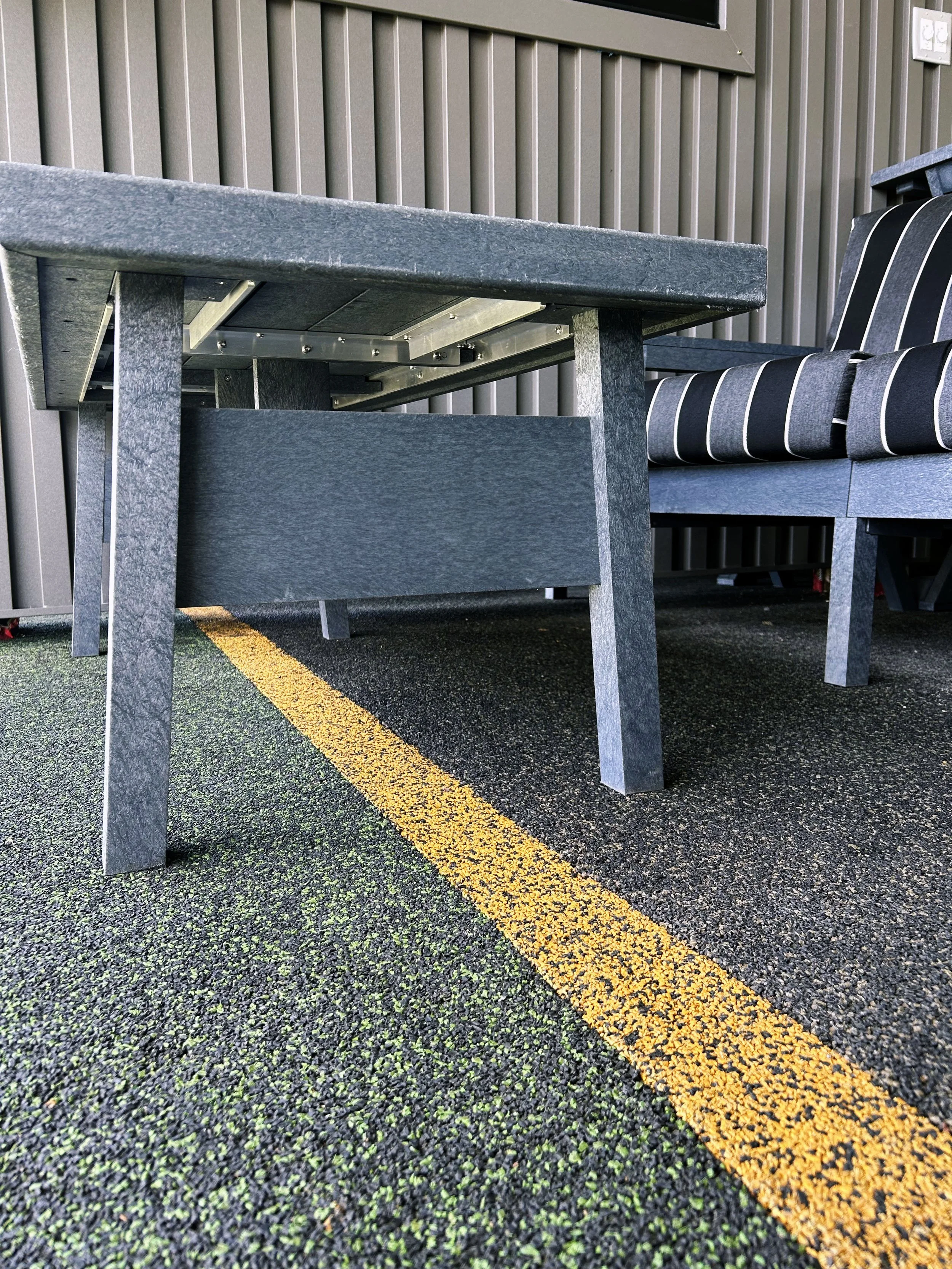 Outdoor seating area with a dark gray stone table and a bench with black and white striped cushions, next to a yellow parking lot line on black asphalt with green flecks.