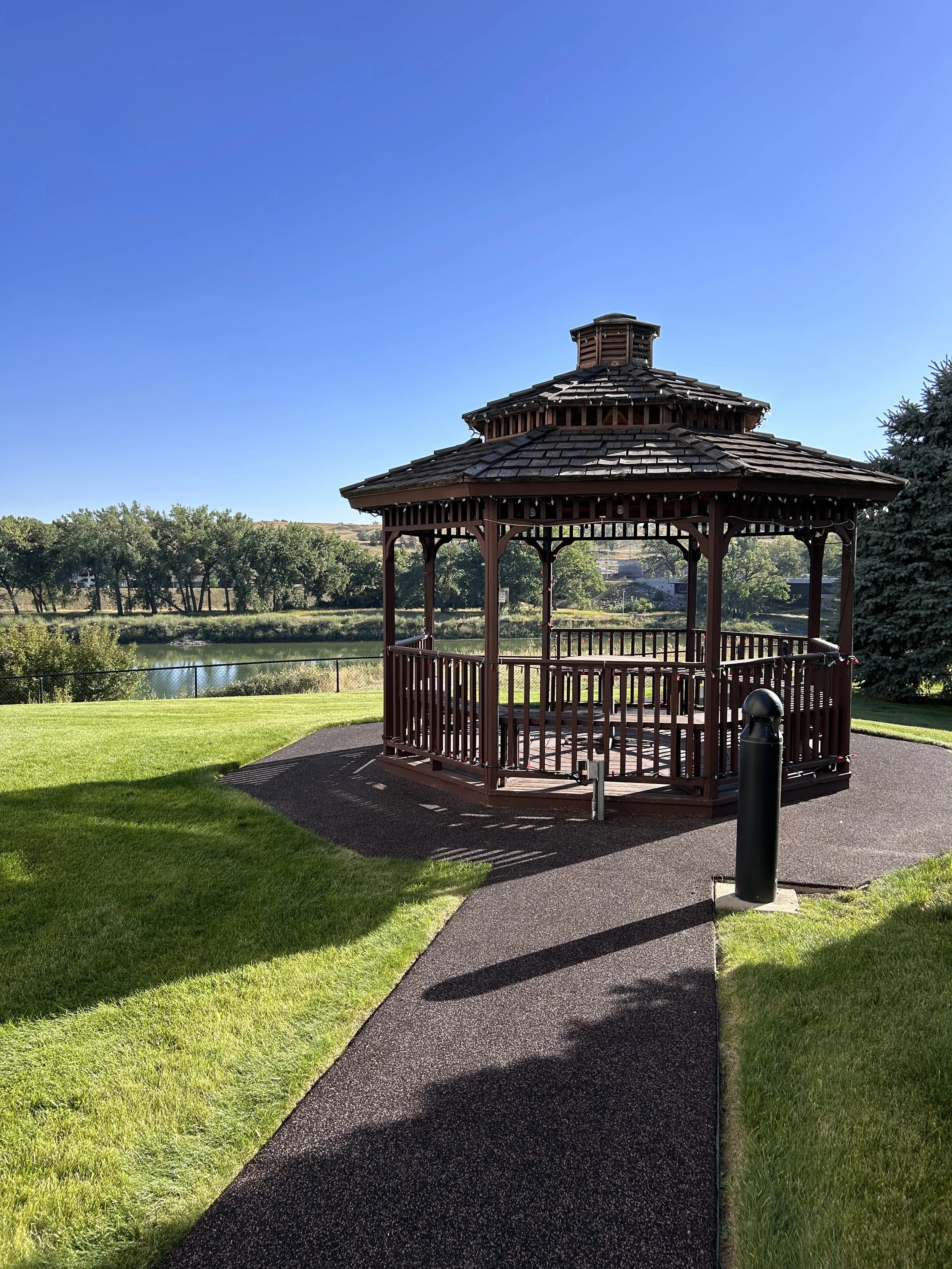 A wooden gazebo with a shingled roof situated in a park with a pond in the background, lush green grass, trees, and a clear blue sky.