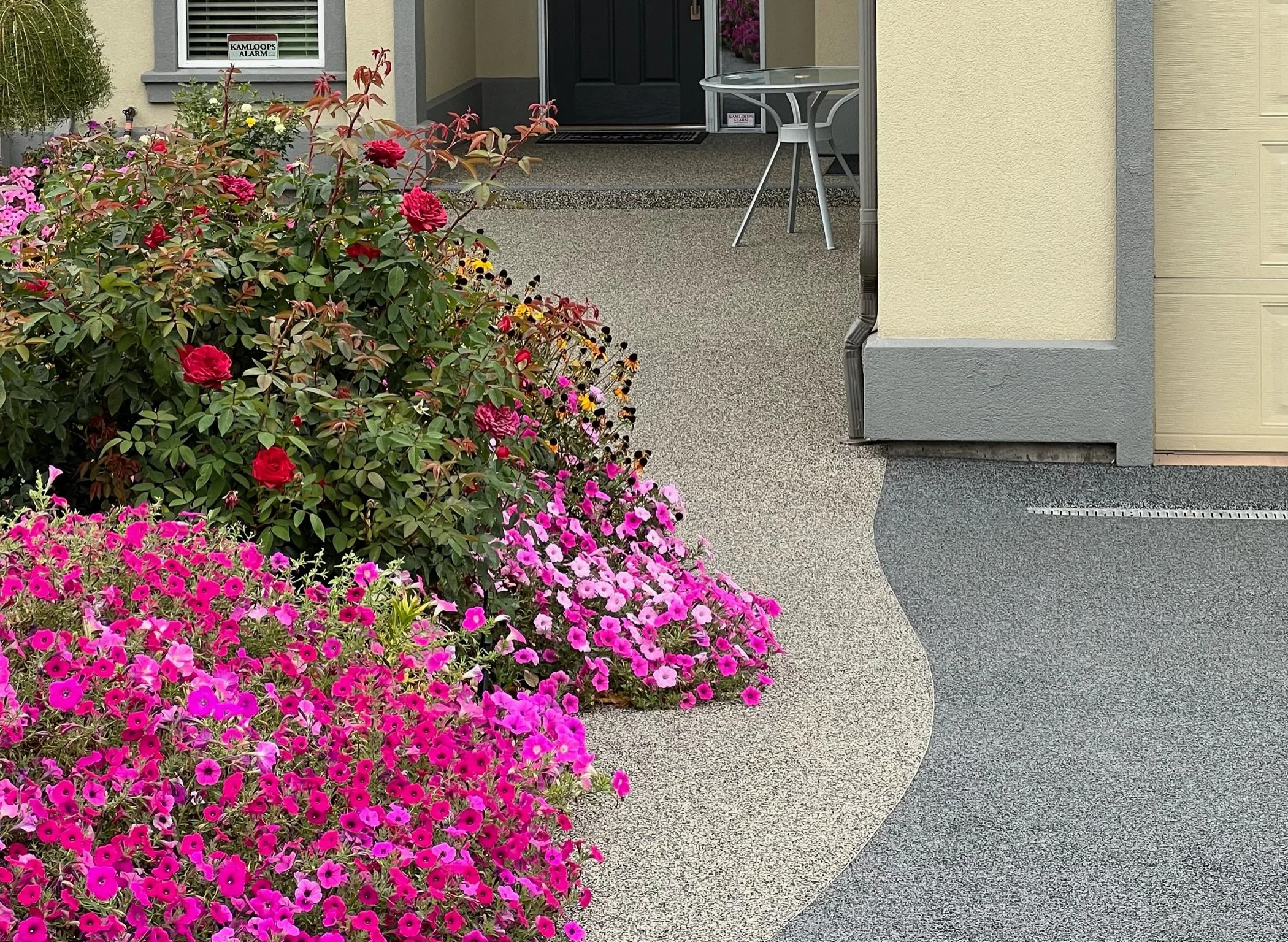 A front porch with a variety of pink, red, and purple flowers in the foreground. The porch has a concrete floor, a small round glass table, and a black front door. There is a window with white blinds and a sign for Kamloops Alarm. Part of a yellow ho