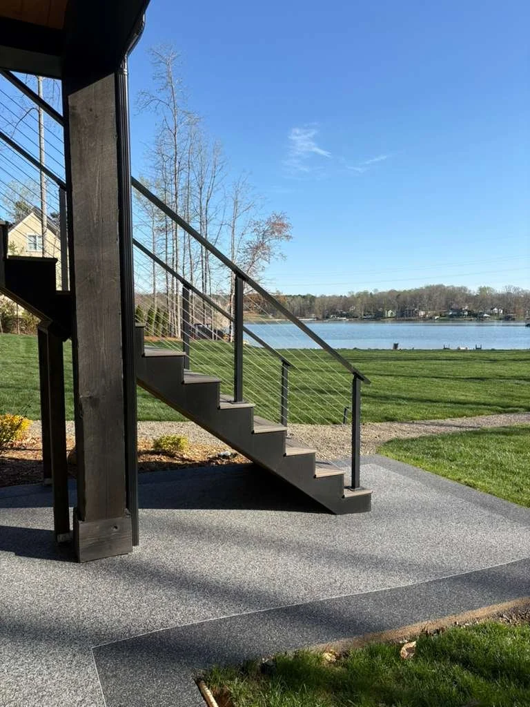 Outdoor view of a staircase leading up to a deck overlooking a grassy area and a lake, under a clear blue sky.