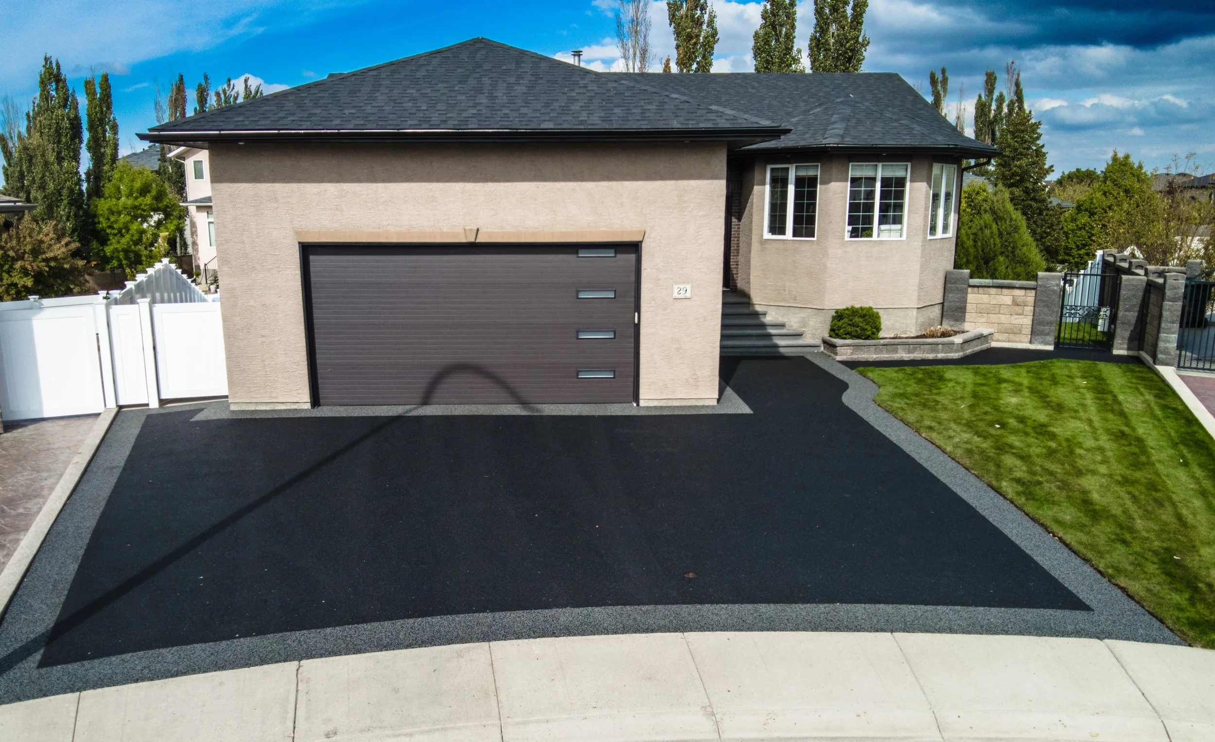 A modern house with a black garage door, beige exterior walls, a bay window on the right, and steps leading to the front door. The front yard has a small lawn and a paved driveway, surrounded by trees and a white fence.