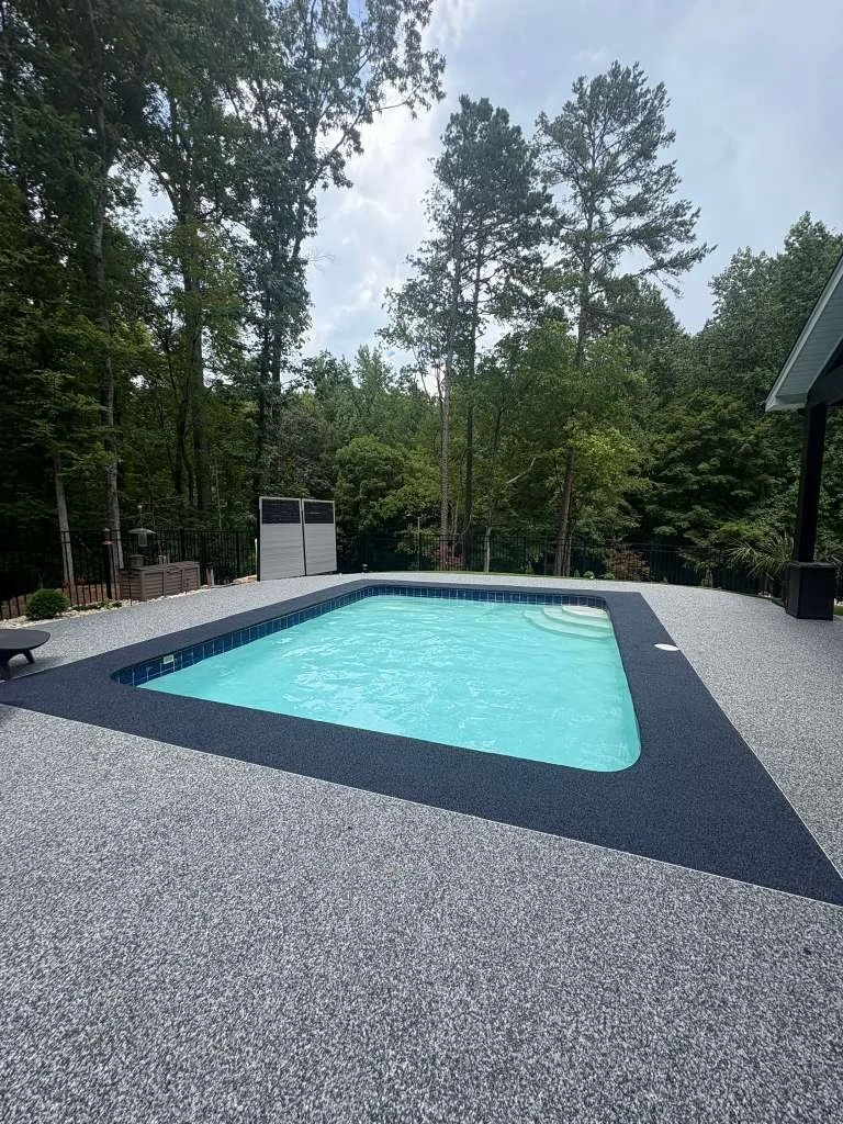 Rectangular backyard swimming pool with a textured grey surround, surrounded by tall trees and a black fence, under a partly cloudy sky.