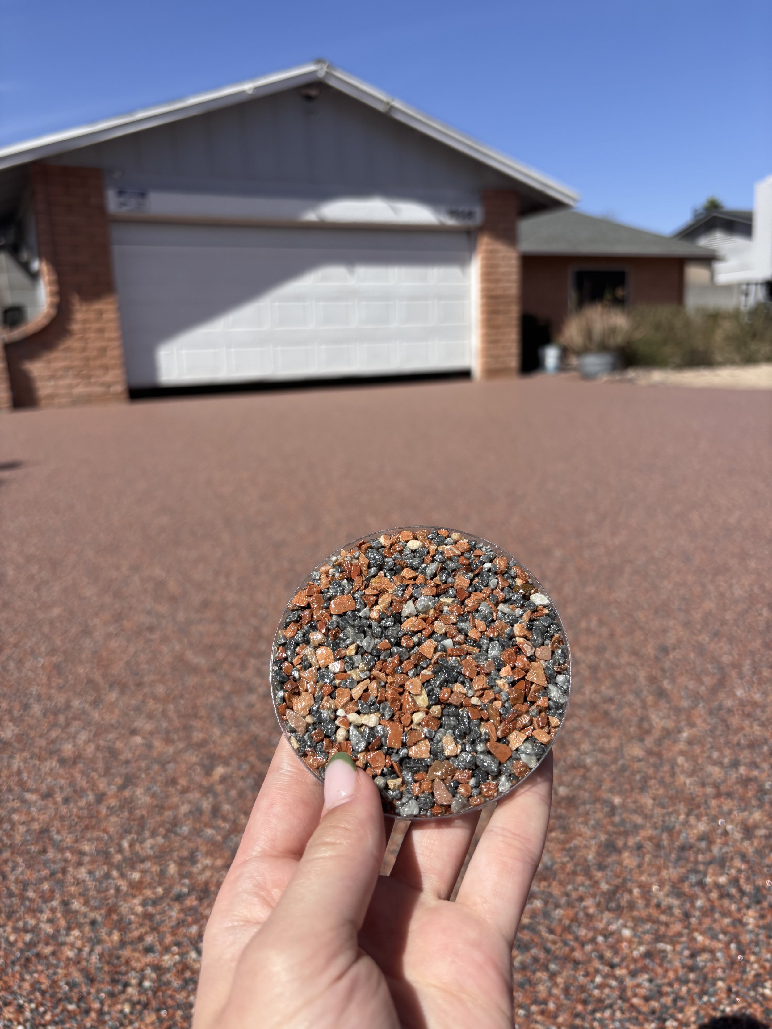 A hand holding a small container of multicolored gravel with a house and a clear blue sky in the background.
