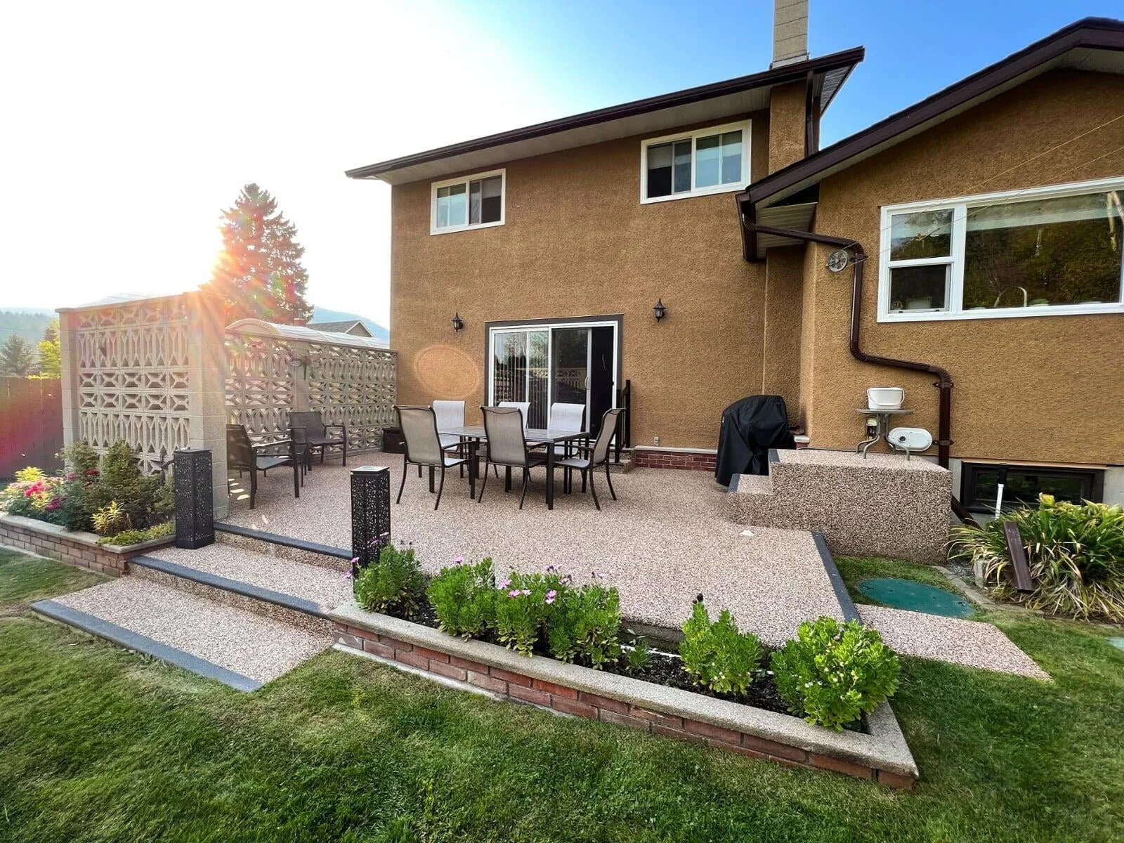 Backyard patio with a dining table and chairs, a barbecue grill, potted plants, and a decorative privacy wall, with the sun setting behind a large tree in the background.