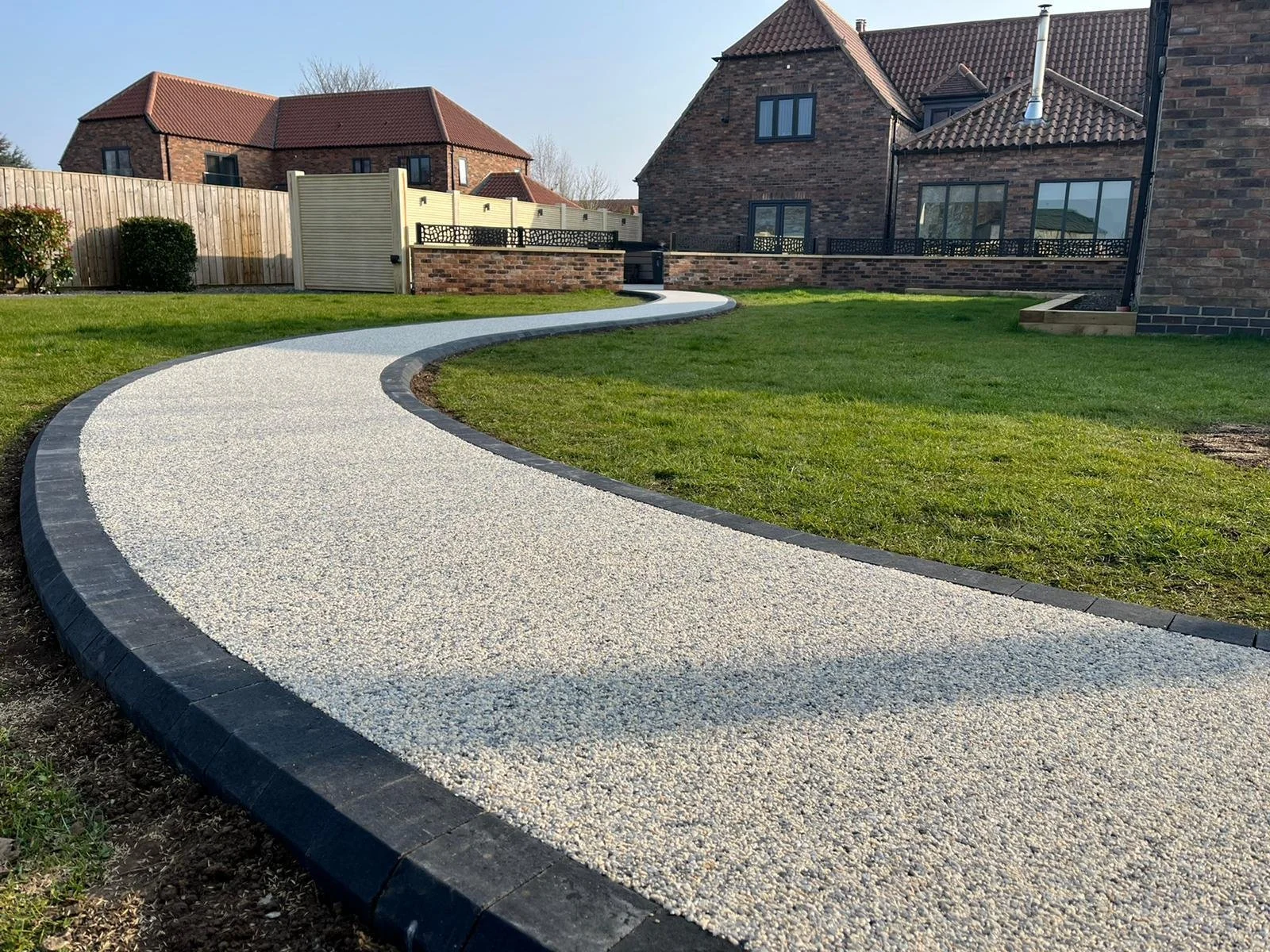 A curved gravel pathway in a backyard yard with green grass, brick houses, and a wooden fence in the background.