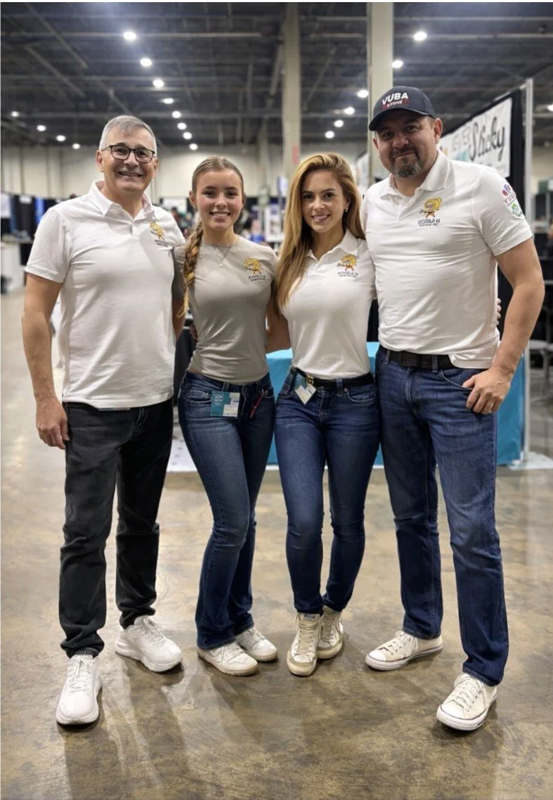 Four people standing together in an indoor setting, all wearing matching white polo shirts with a logo featuring a lion and the text 'Coca-Cola U,' and jeans. They are smiling and have their arms around each other.