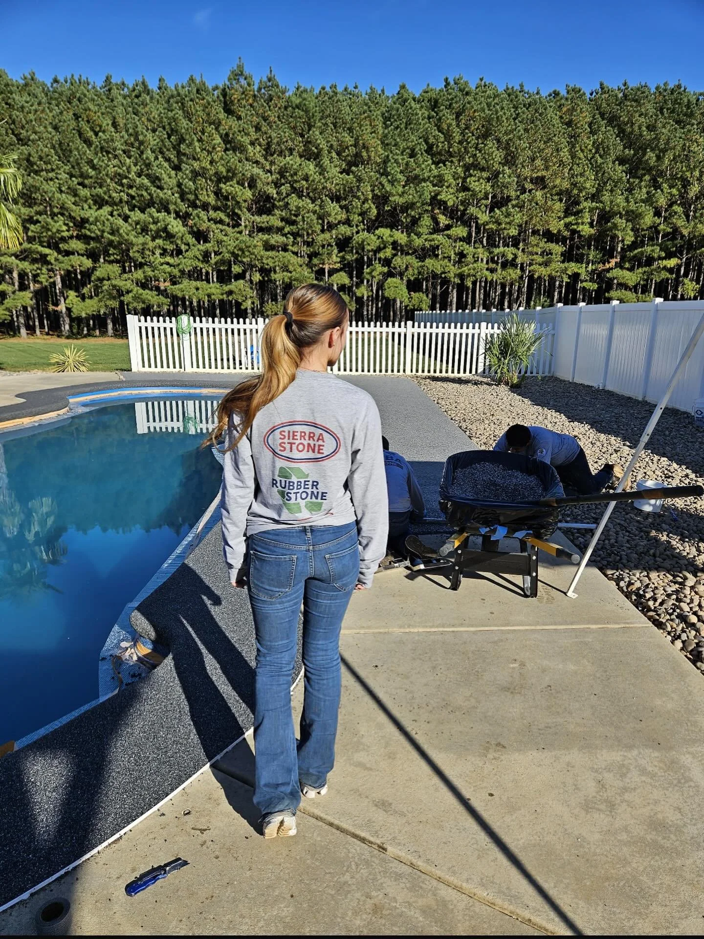 A woman with long hair in a ponytail wearing a gray Sierra Stone sweatshirt and blue jeans standing on a concrete patio next to a backyard pool. Two workers are kneeling beside a black container, working on the ground. The yard is enclosed by a white