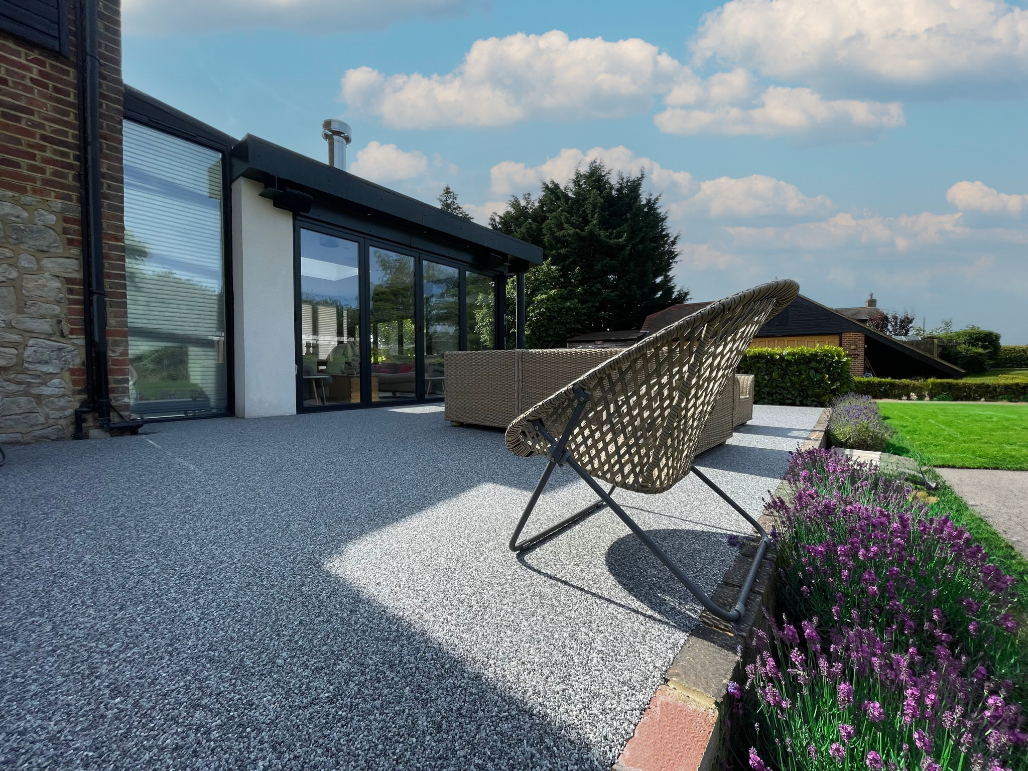 A modern house with a large glass door and a patio area with outdoor furniture, surrounded by a well-kept garden with purple flowers, next to a brick and stone exterior wall under a partly cloudy sky.